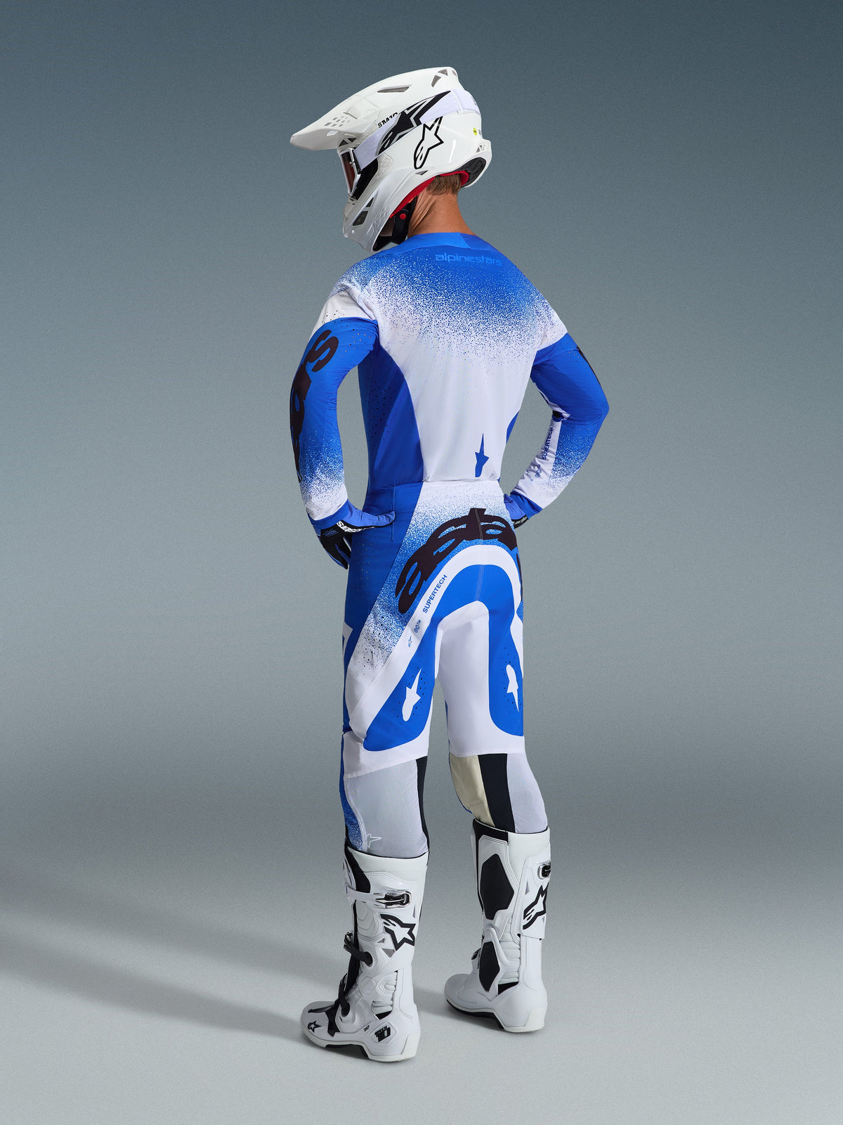 Motocrossförare sedd bakifrån klädd i 2026 Supertech Scenz-byxor och tröja i Ucla Blue White, vit hjälm med Alpinestars-logotyp, vita och svarta motocross-stövlar, professionell racingutrustning för terrängkörning.