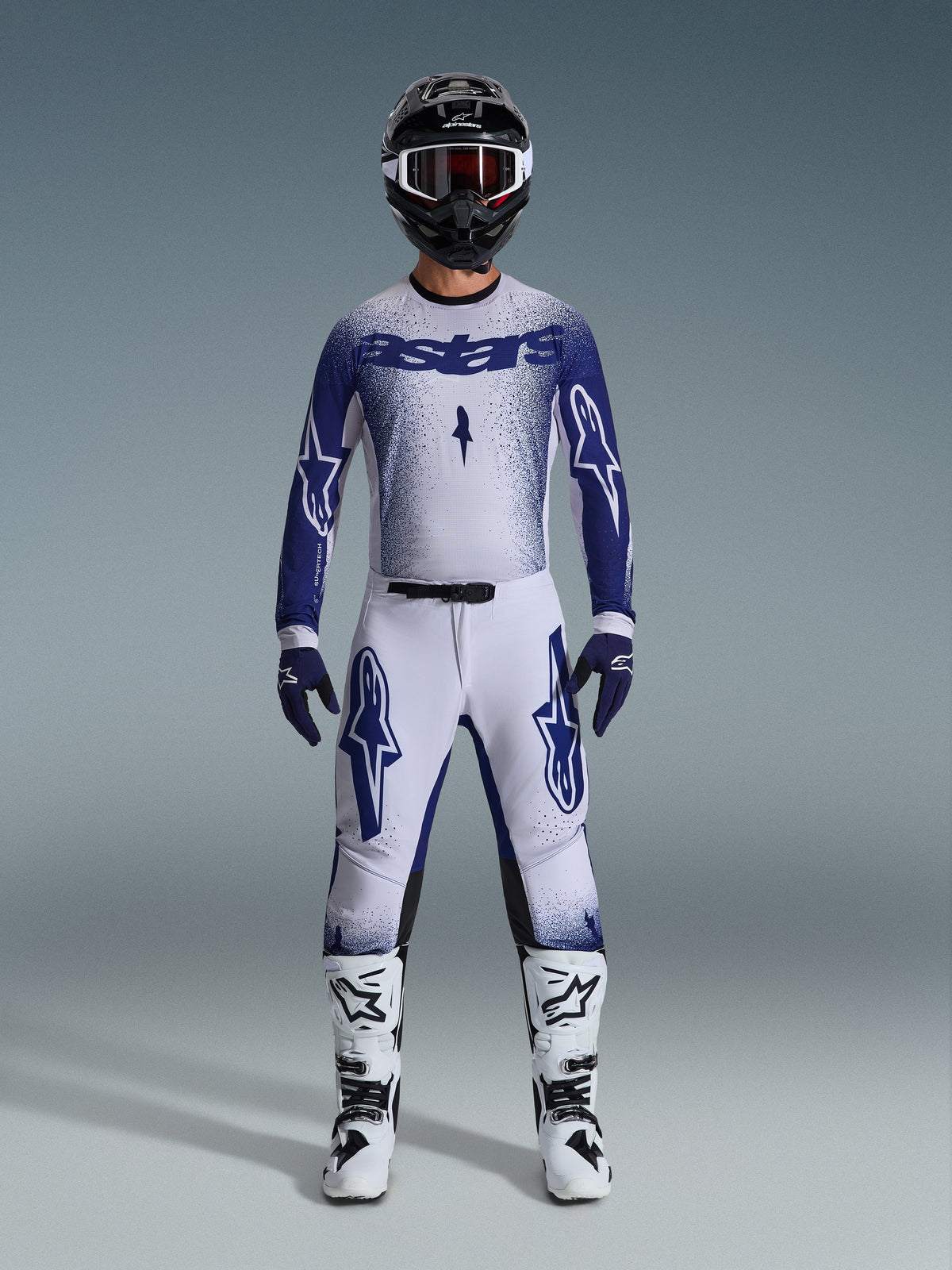 Motocross rider wearing Alpinestars 2026 Supertech Scenz gear set, Light Gray and Dark Blue, including a jersey and pants with matching blue gloves, white and black boots, and a black helmet with white goggles, coordinated outfit for professional off-road racing