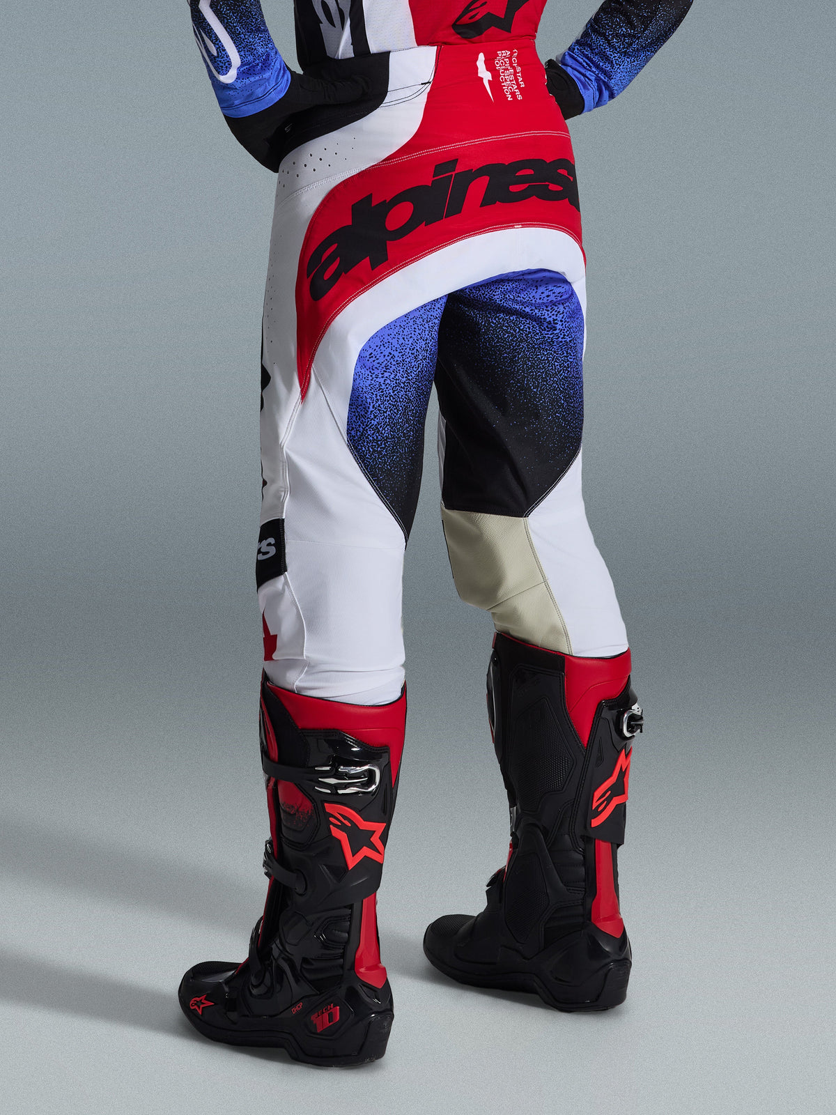 Alpinestars 2026 Techstar Knif Pants, Motocross Pants, White Red and Blue, back view showing red seat with black branding, blue and black speckled patterns on legs, worn with black and red Tech 10 boots, elite off-road racing gear