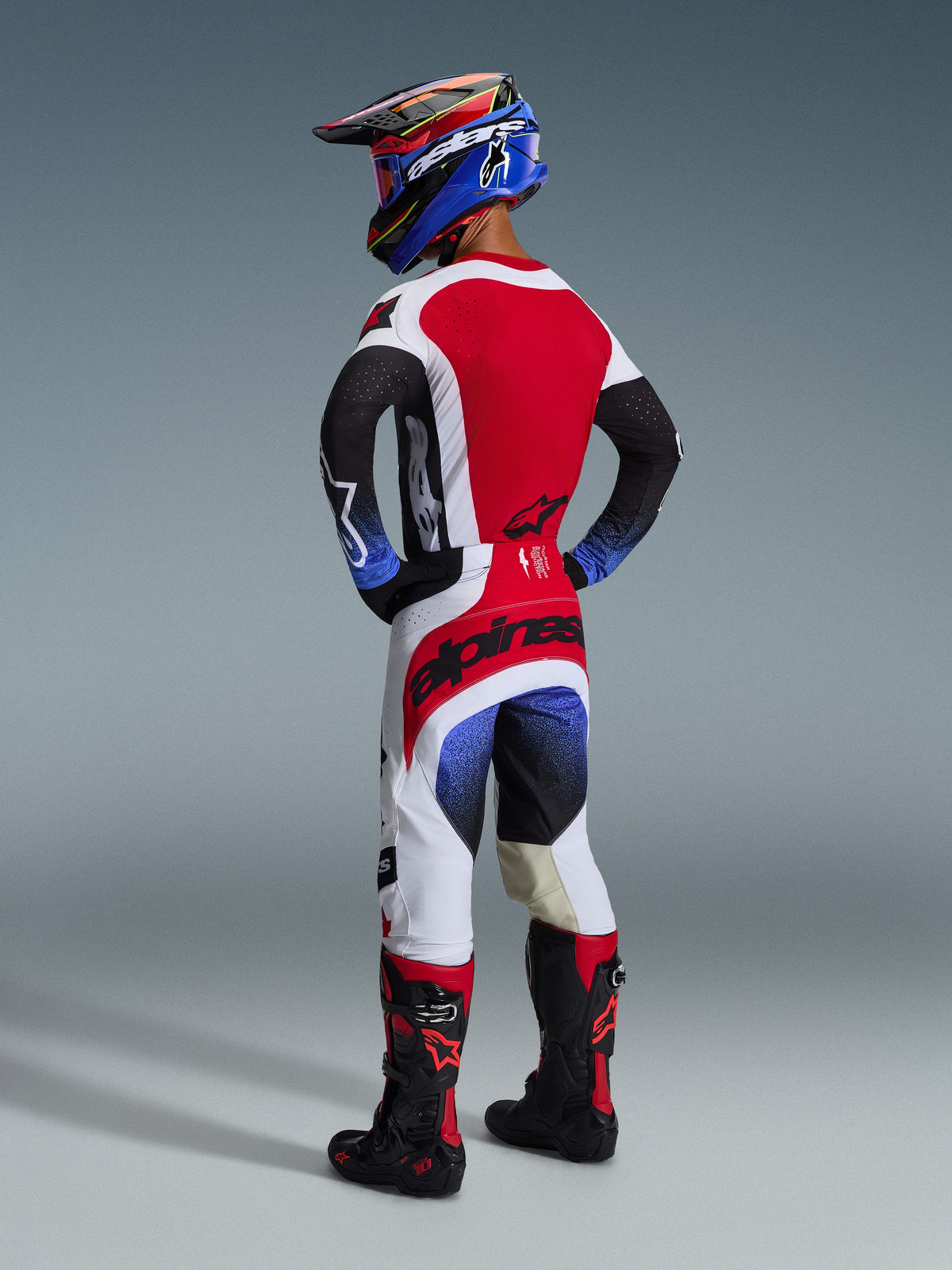 Alpinestars 2026 Techstar Knif Pants, Motocross Pants, Black White and Red with blue gradient accents, back view of rider wearing full racing gear, bold red branding on the seat, white and black paneling, worn with matching jersey and Tech 10 boots.