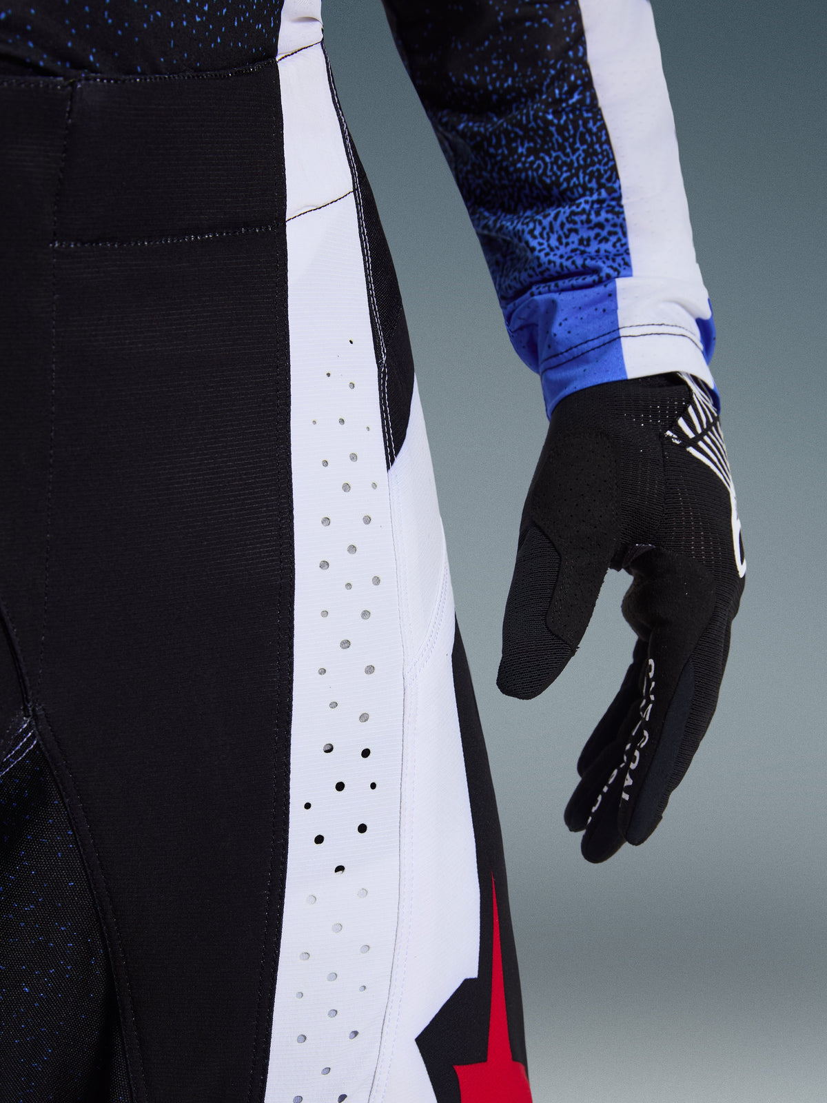 Close-up detail of Alpinestars 2026 Techstar Knif Pants in Black White and Red, showcasing laser-cut perforations on a white side panel for ventilation, shown with a black motocross glove and blue patterned jersey sleeve, highlighting high-performance racing construction.