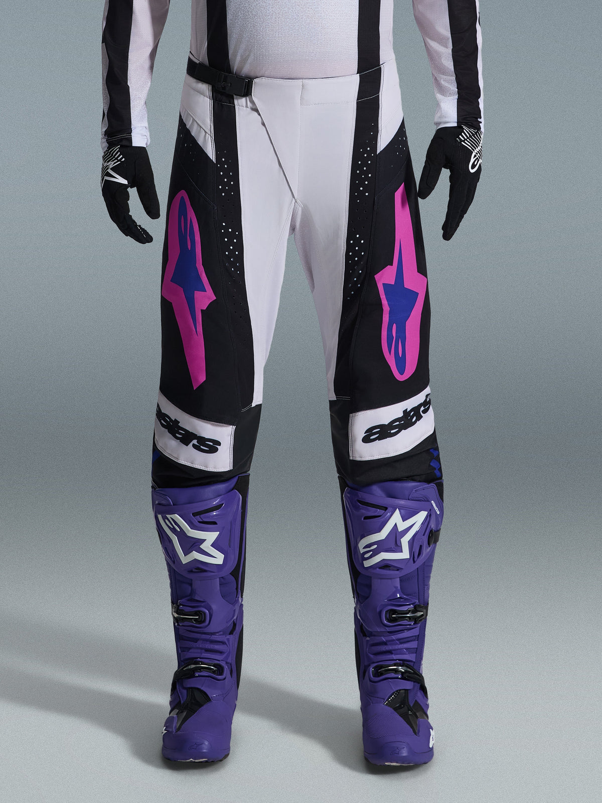 Motocrossförare i Alpinestars 2026 Techstar Knif-utrustningsset, vita och svarta byxor med rosa och blå logotyper, lila stövlar med vit branding, svarta handskar, atletisk racingpassform och laserskuren ventilation för terrängkörning