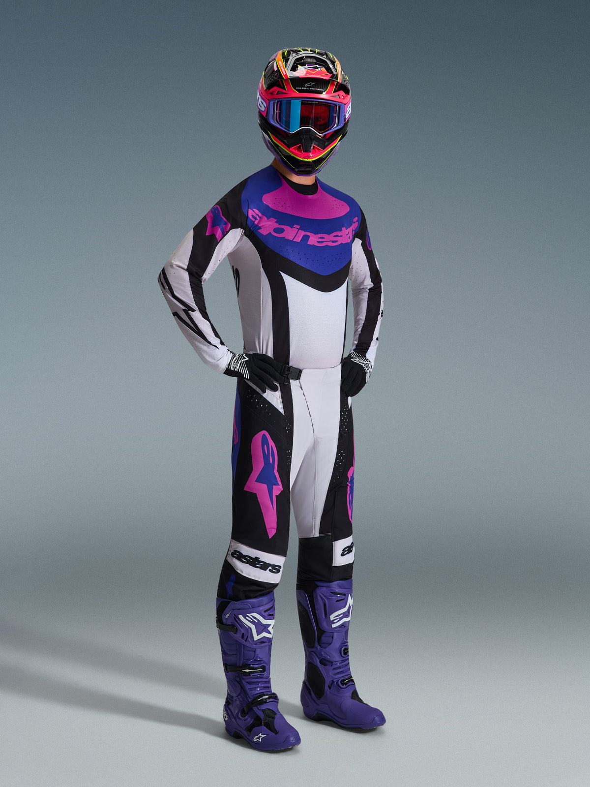 Motocrossförare i Alpinestars 2026 Techstar Knif-utrustningsset, vit tröja med svarta, blå och rosa detaljer, matchande byxor med rosa Astars-logotyper, flerfärgad hjälm med goggles, lila racingstövlar, koordinerad högpresterande utrustning för motocrosstävling