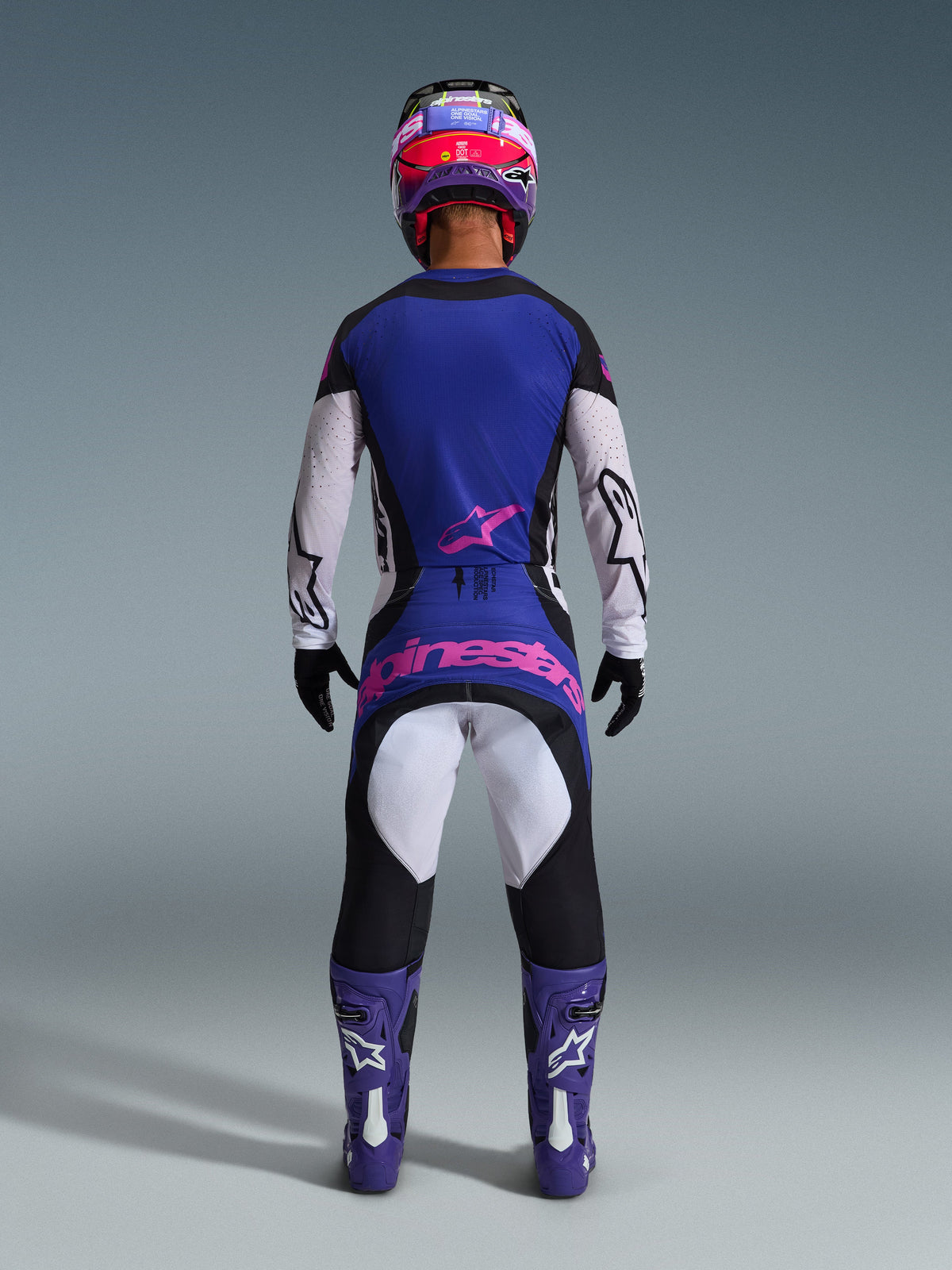 Vy bakifrån av en motocrossförare i Alpinestars 2026 Techstar Knif-utrustningsset, blå och vit tröja med rosa stjärnlogotyp, matchande blå och svarta byxor med stor rosa Alpinestars-logotyp på baksidan, lila och vita stövlar samt en lila och röd hjälm, koordinerad professionell racingoutfit för terrängkörning