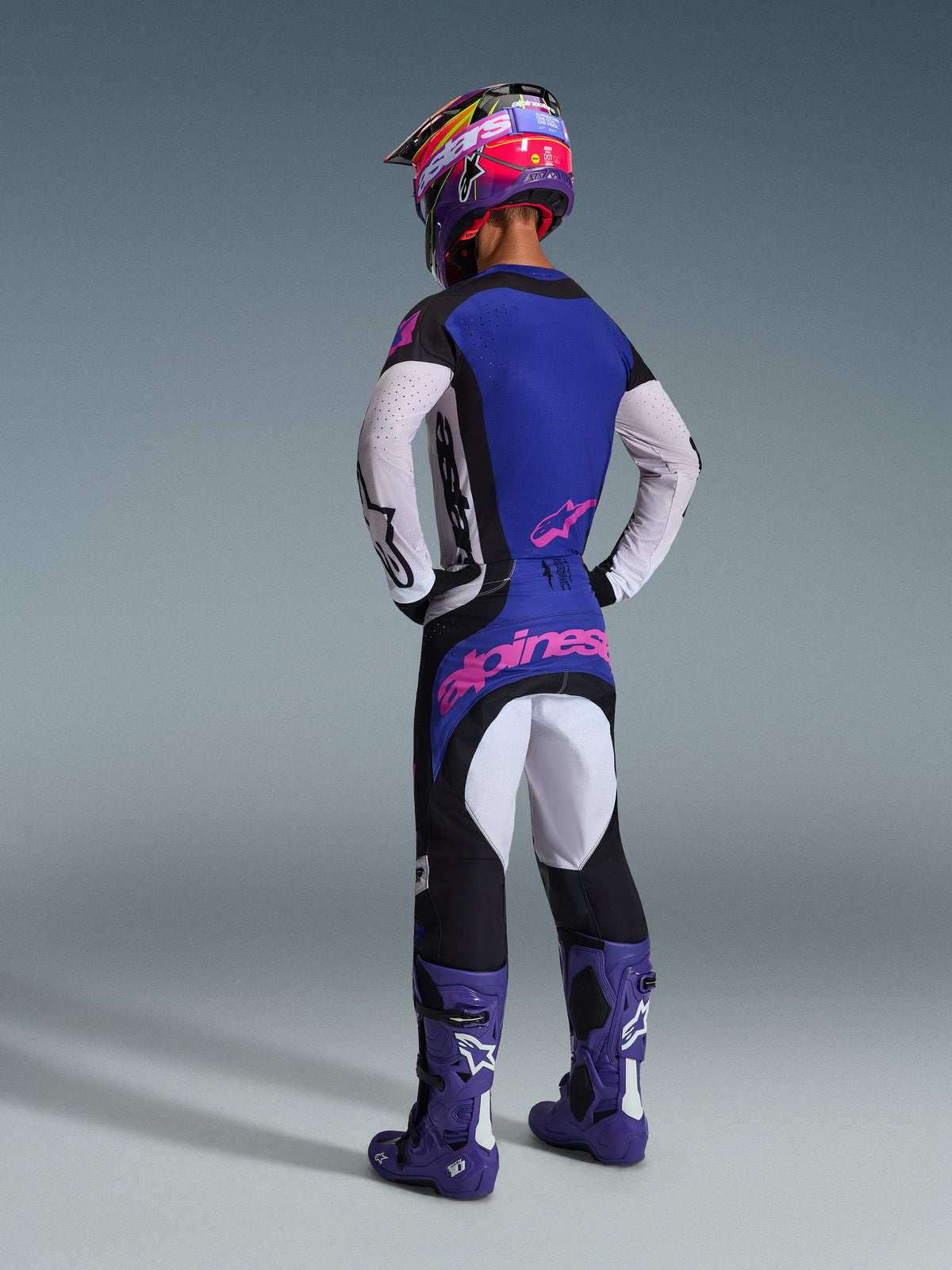 Motocrossförare sedd bakifrån i Alpinestars 2026 Techstar Knif utrustningsset, blå tröja med vita perforerade ärmar och rosa logotyper, svarta och vita byxor med rosa logotyp på baksidan, matchat med lila stövlar och en livfull lila och rosa hjälm, professionell motocrossoutfit.