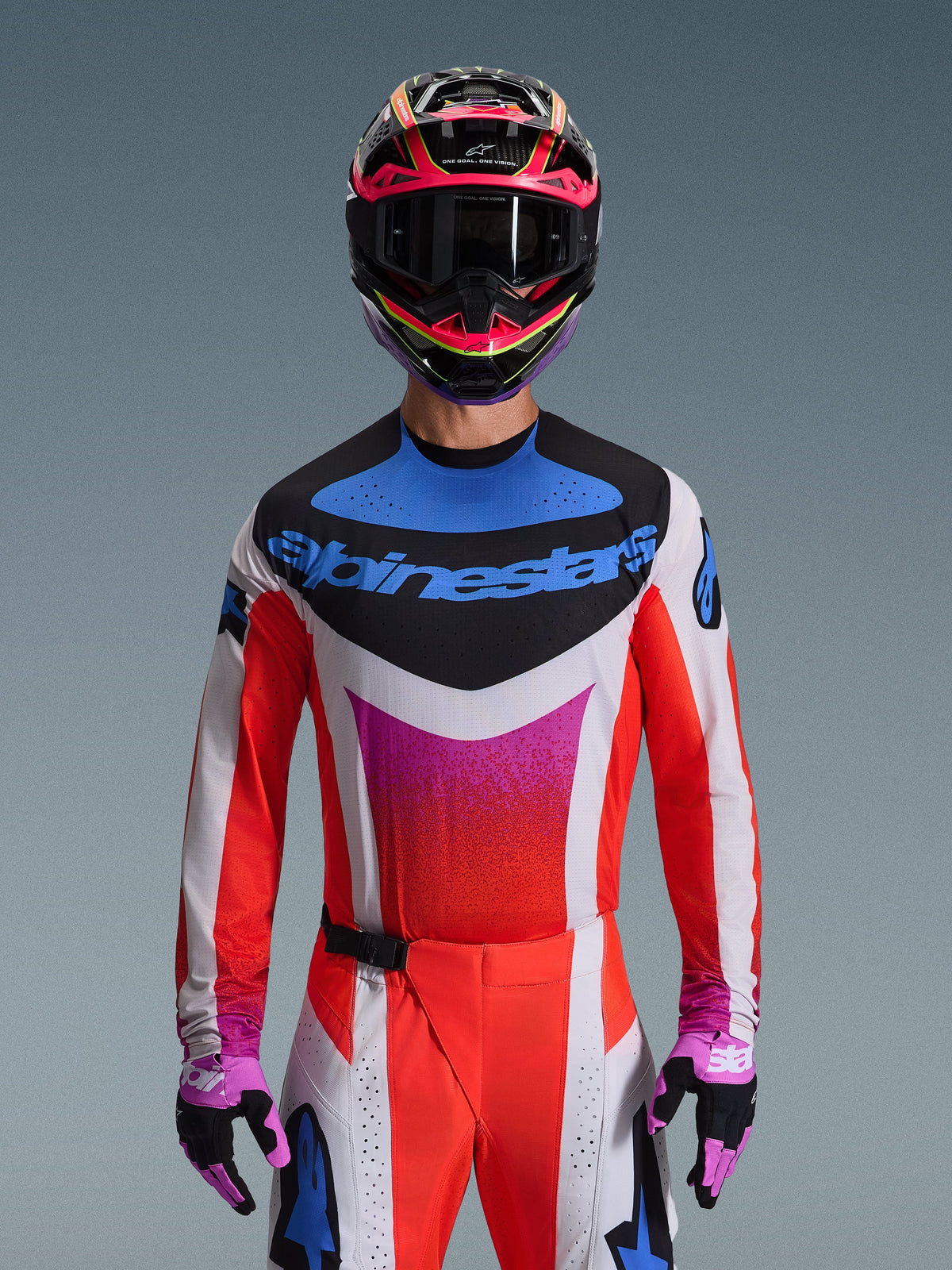 Motocrossförare klädd i 2026 Techstar Knif-utrustningsset, vit tröja med röda ärmar och blå och rosa grafik på bröstet, matchande röda och vita byxor, färgstark rosa och gul hjälm med mörka goggles och lila och svarta handskar, professionell utrustning för terrängkörning.