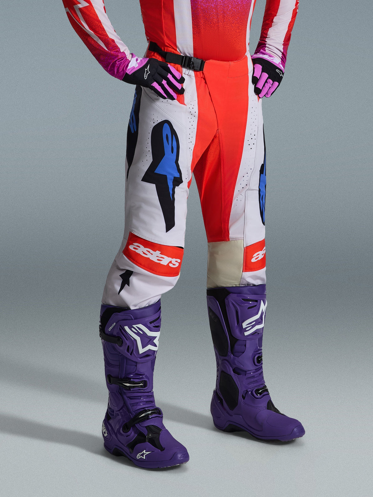 Alpinestars 2026 Techstar Knif-byxor, motocrossbyxor, vita och orangea med blå detaljer, bärs som en del av ett komplett utrustningsset med röd och rosa tröja, lila stövlar och rosa handskar, framträdande logotyper och atletisk racingpassform