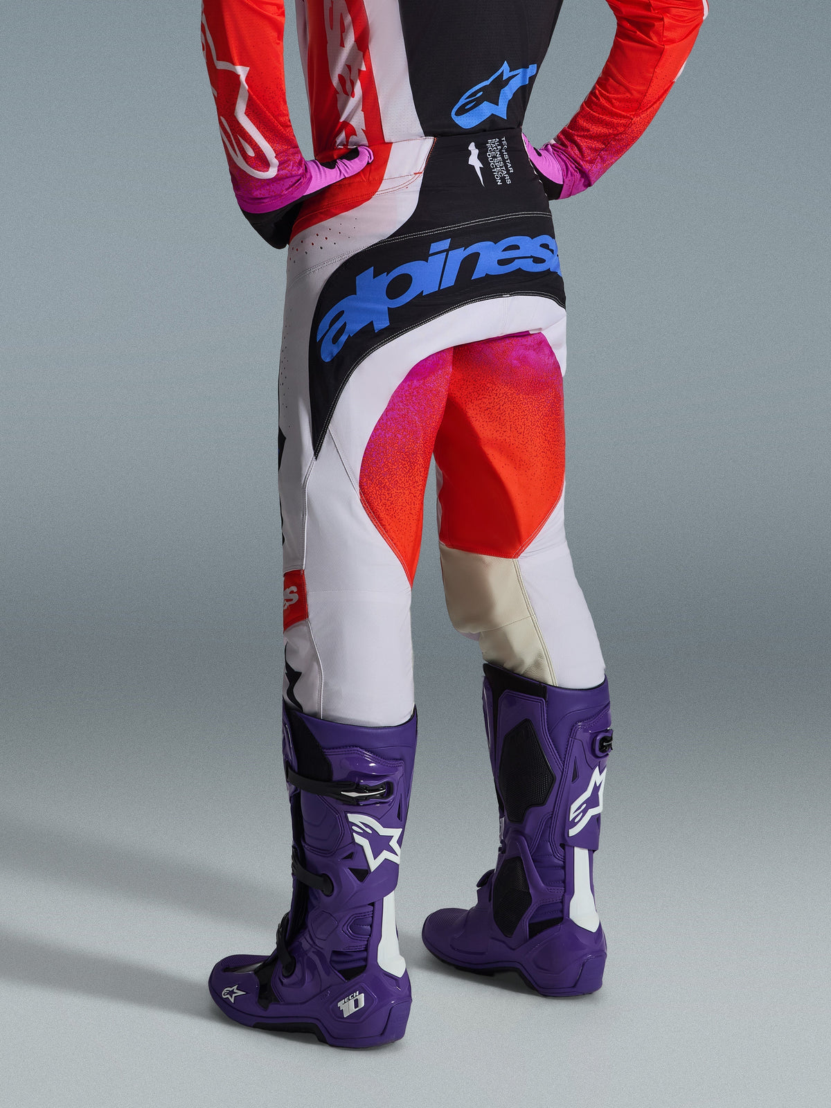 Alpinestars 2026 Techstar Knif byxor och utrustningsset, vy bakifrån av en motocrossförare i vita byxor med svarta paneler, rosa och röda tonade accenter och en stor blå Alpinestars-logotyp på sätet, matchat med lila Tech 10-stövlar och en röd och svart tröja.