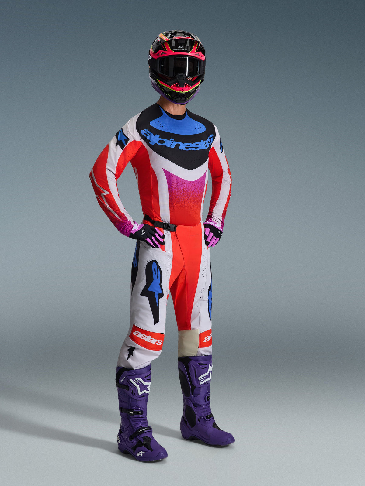 Alpinestars 2026 Techstar Knif utrustningsset, motocrossförare i komplett racingutrustning, röd tröja med vita och blå detaljer, vita byxor med röda och blå stjärnlogotyper, lila motocross-stövlar, svart och rosa hjälm, professionellt kit för terrängkörning