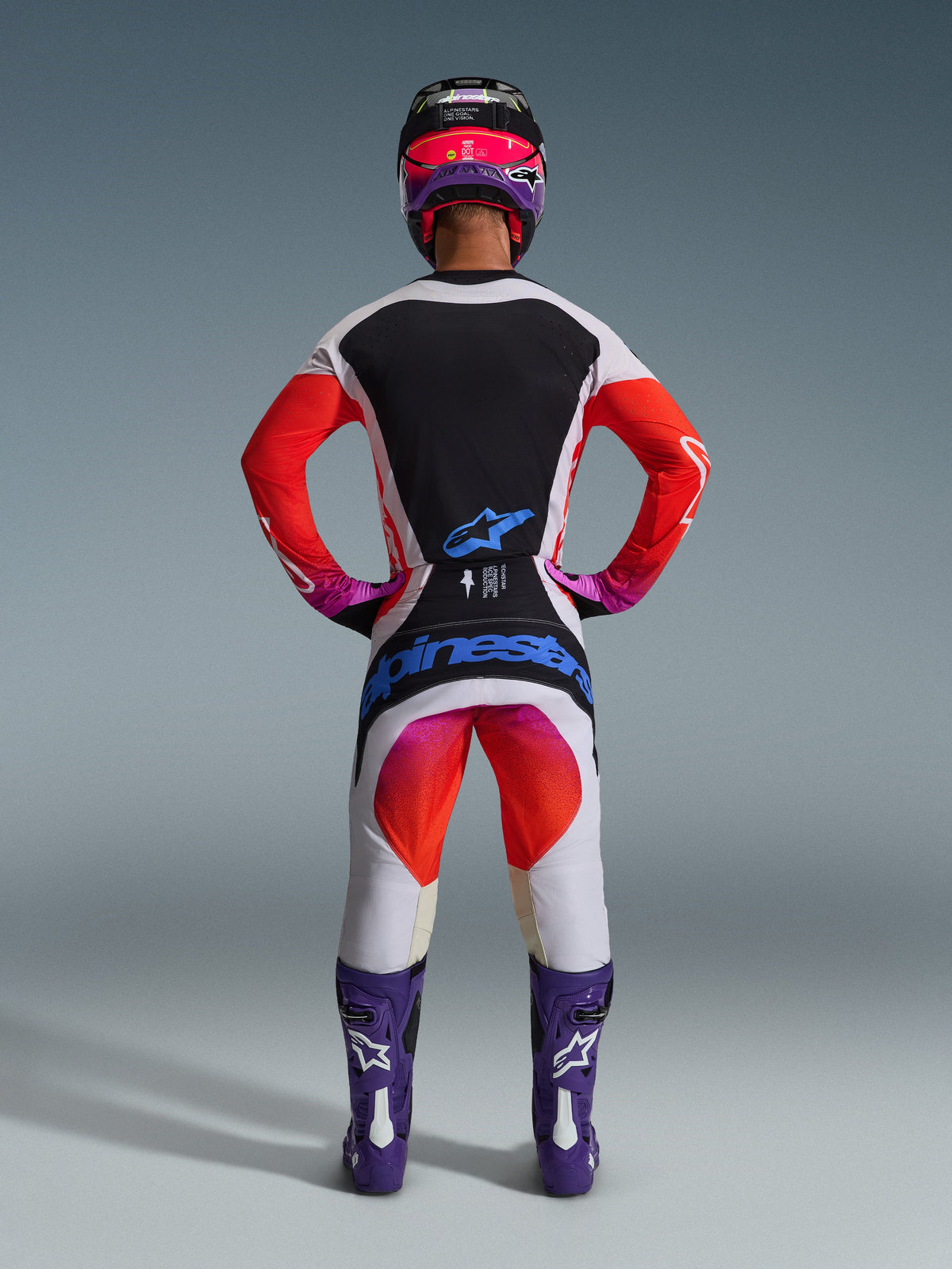 Motocrossförare som bär komplett 2026 Techstar Knif racingutrustning sedd bakifrån, ljusgrå tröja med svart ryggpanel och orange ärmar, matchande vita och orangea byxor med blå Alpinestars-logotyper, lila stövlar och en svart och rosa hjälm, koordinerad professionell outfit för terrängkörning
