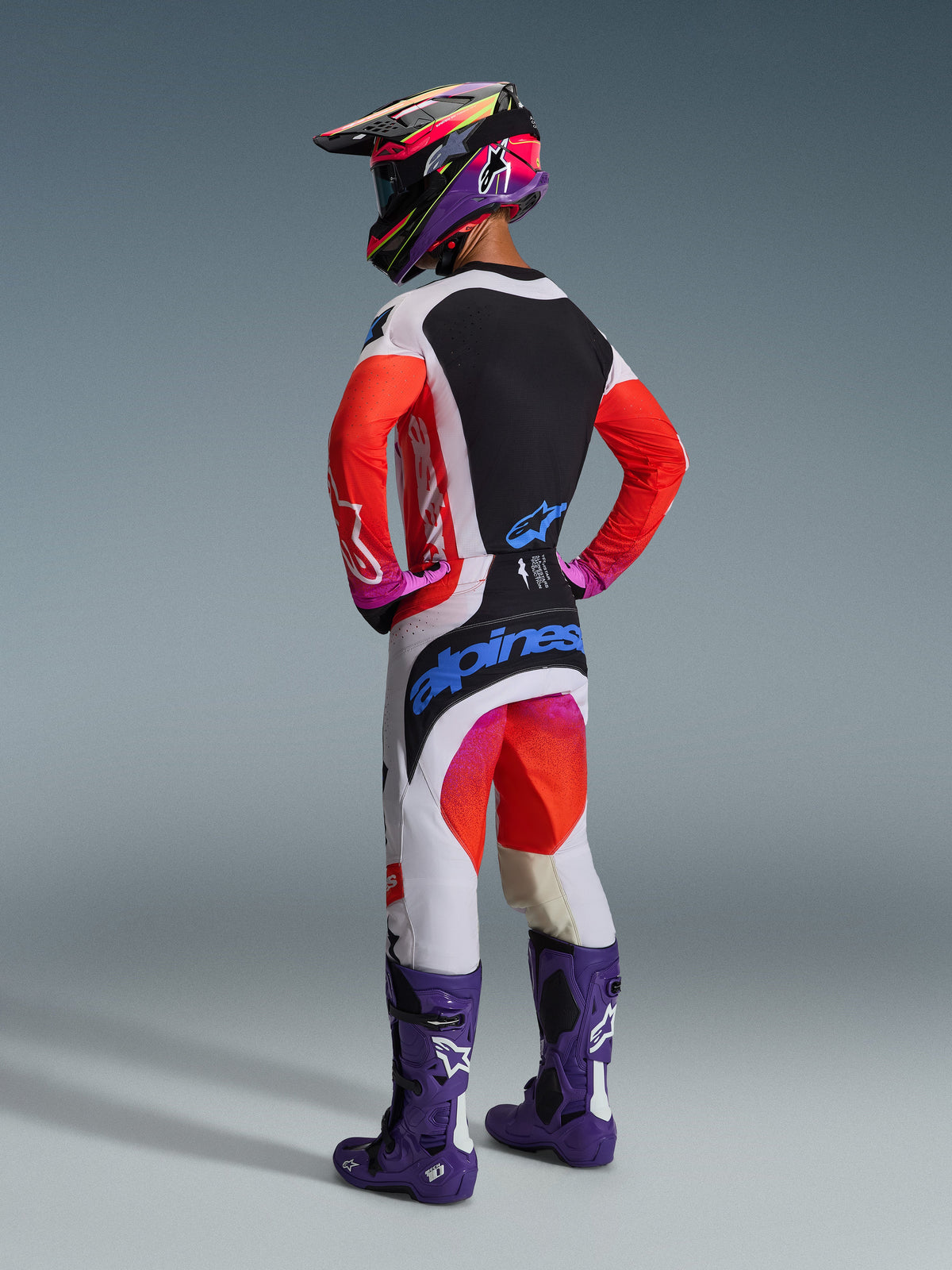 Motocrossförare sedd bakifrån i Alpinestars 2026 Techstar Knif-utrustningsset, vit tröja med röda ärmar och svart panel, vita byxor med röda och lila detaljer och blå Alpinestars-logotyp, lila racingstövlar och matchande hjälm, professionell utrustning för terrängkörning