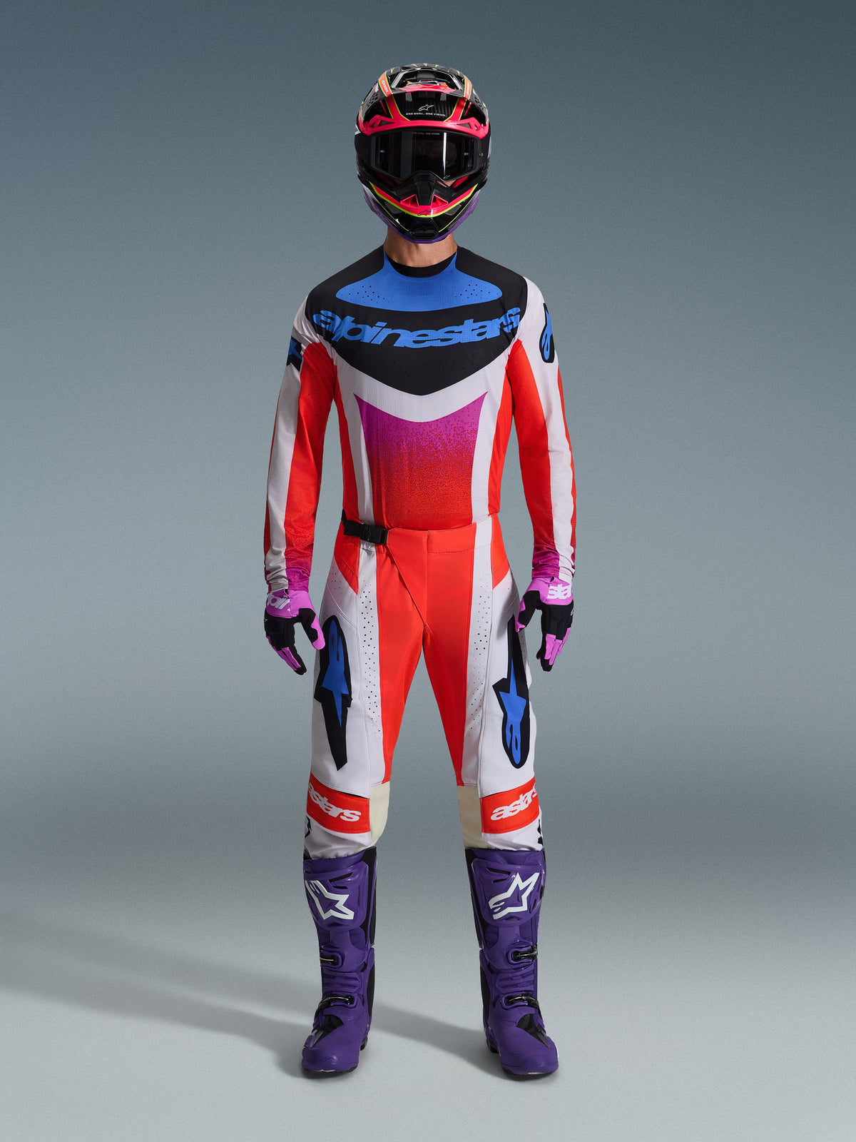 Motocrossförare i komplett racingutrustning, Alpinestars 2026 Techstar Knif-tröja och matchande byxor i vitt och orange med blå och lila detaljer, lila racingstövlar med vit logotyp och en flerfärgad hjälm, koordinerad outfit för terrängracing