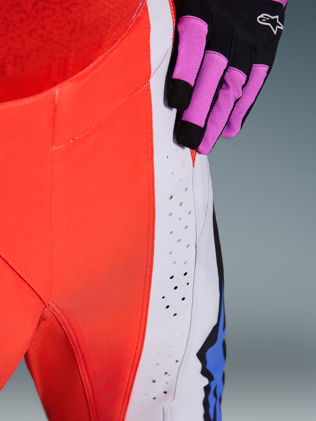 Alpinestars 2026 Techstar Knif byxor, närbild av höft och sidopanel i Orange Fluo och ljusgrått, med laserskurna perforeringar för luftflöde, bärs med en svart och lila motocrosshandske, högpresterande racingutrustning