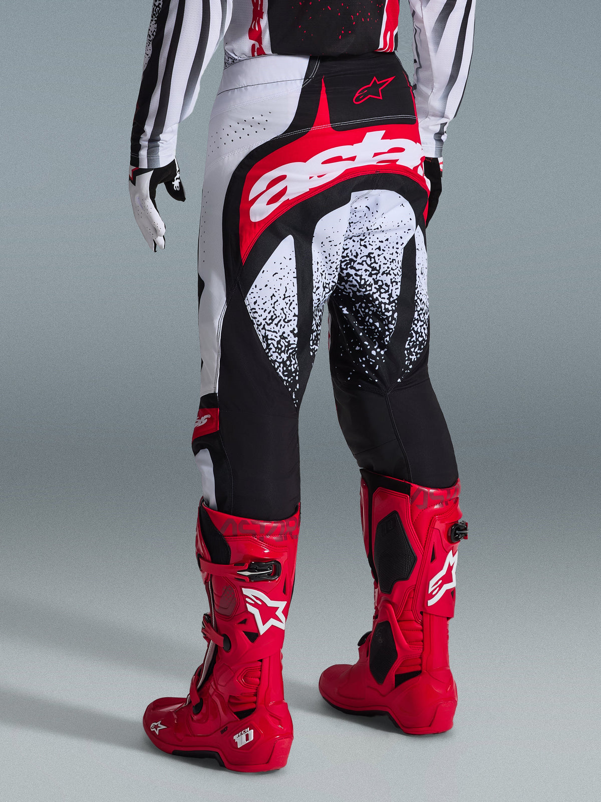 Alpinestars 2026 Techstar Nomur-byxor, motocrossbyxor, svarta, vita och röda, vy bakifrån på en förare med matchande röda stövlar, med en framträdande röd och vit 'astars'-logotyp på sätet och spräckliga grafiska mönster, professionell utrustning för terrängkörning