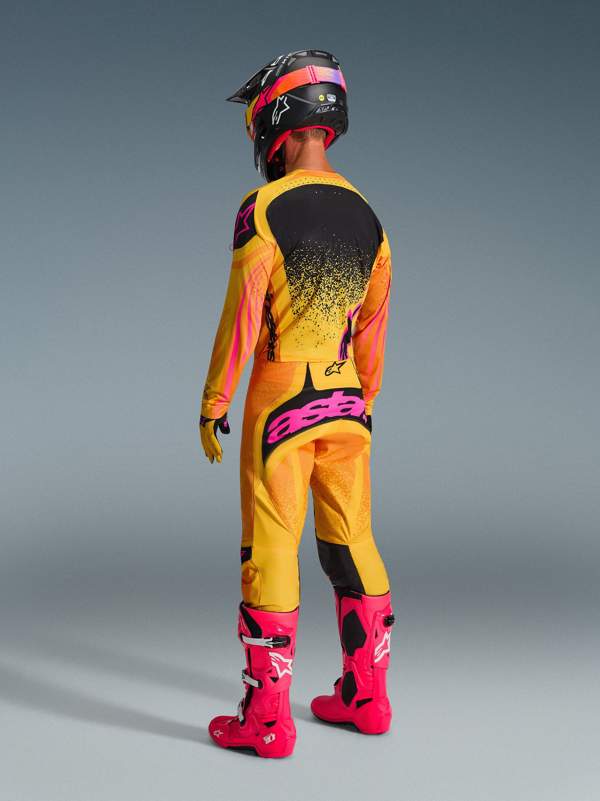Motocross rider viewed from the back wearing complete racing gear, 2026 Techstar Nomur Pants in Orange Yellow Pink, bold pink 'astars' branding on the seat, matching jersey, bright pink boots, and black helmet with pink accents, coordinated off-road competition outfit.