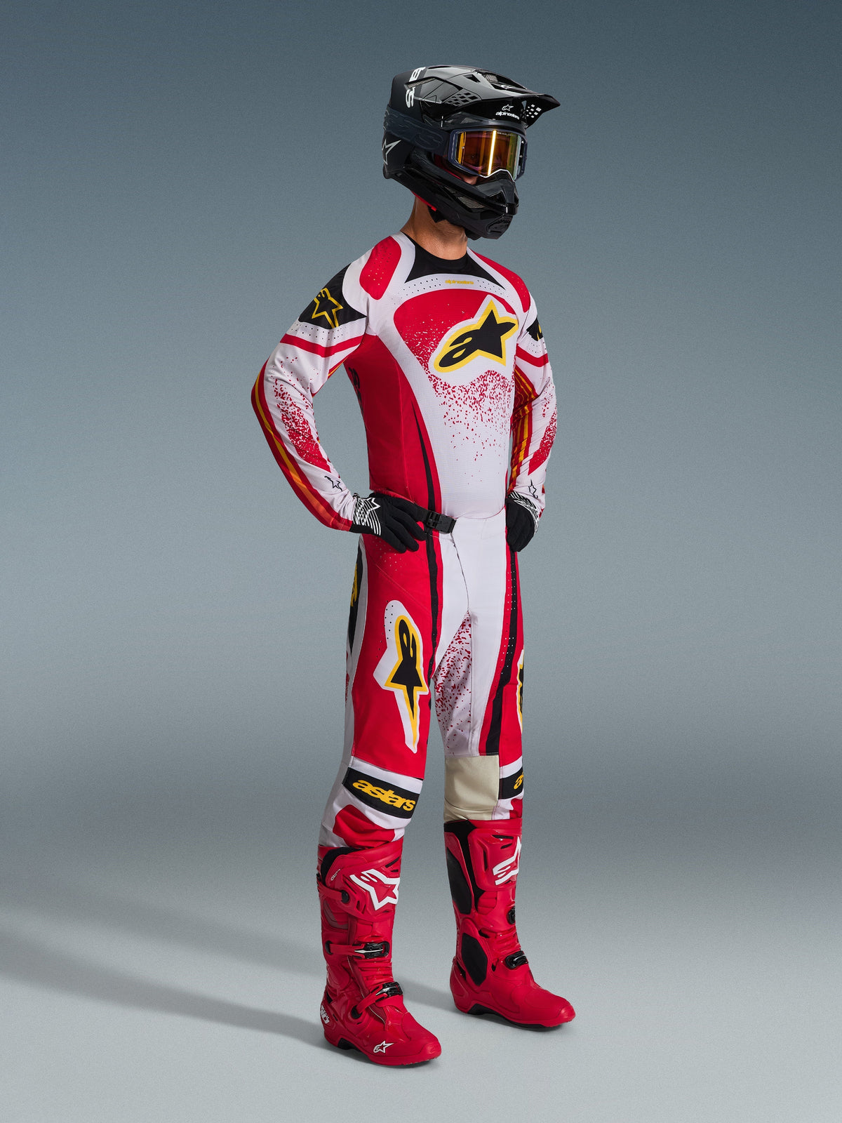 Alpinestars 2026 Techstar Nomur Pants, Motocross Pants, Red and White with black and yellow accents, worn by a rider with a matching racing jersey, bright red boots, and a black helmet with gold goggles, complete professional off-road competition outfit