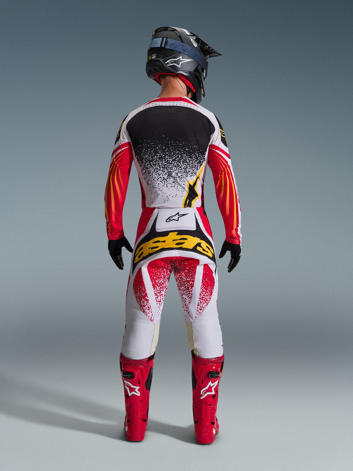 Motocross rider from behind wearing complete racing gear, black and white jersey with red sleeves and yellow accents, 2026 Techstar Nomur Pants in white and red with a large yellow 'astars' logo on the back, bright red boots, and black helmet, coordinated outfit for off-road racing