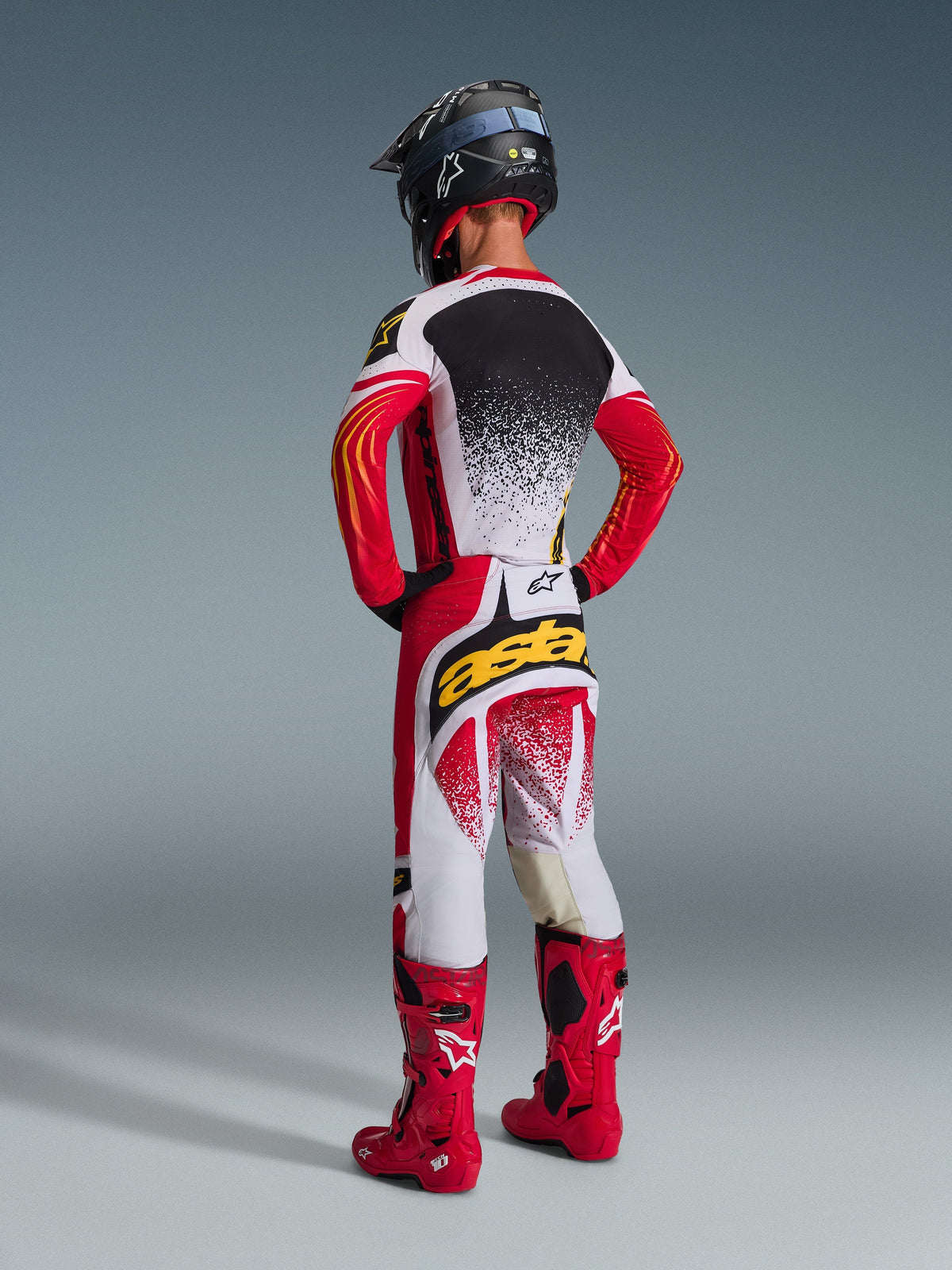 Motocross rider from behind wearing complete Alpinestars racing gear, 2026 Techstar Nomur Pants in Red White and Black, matching jersey with speckled design, bright red boots, black helmet, bold 'astars' branding on seat, coordinated off-road racing outfit