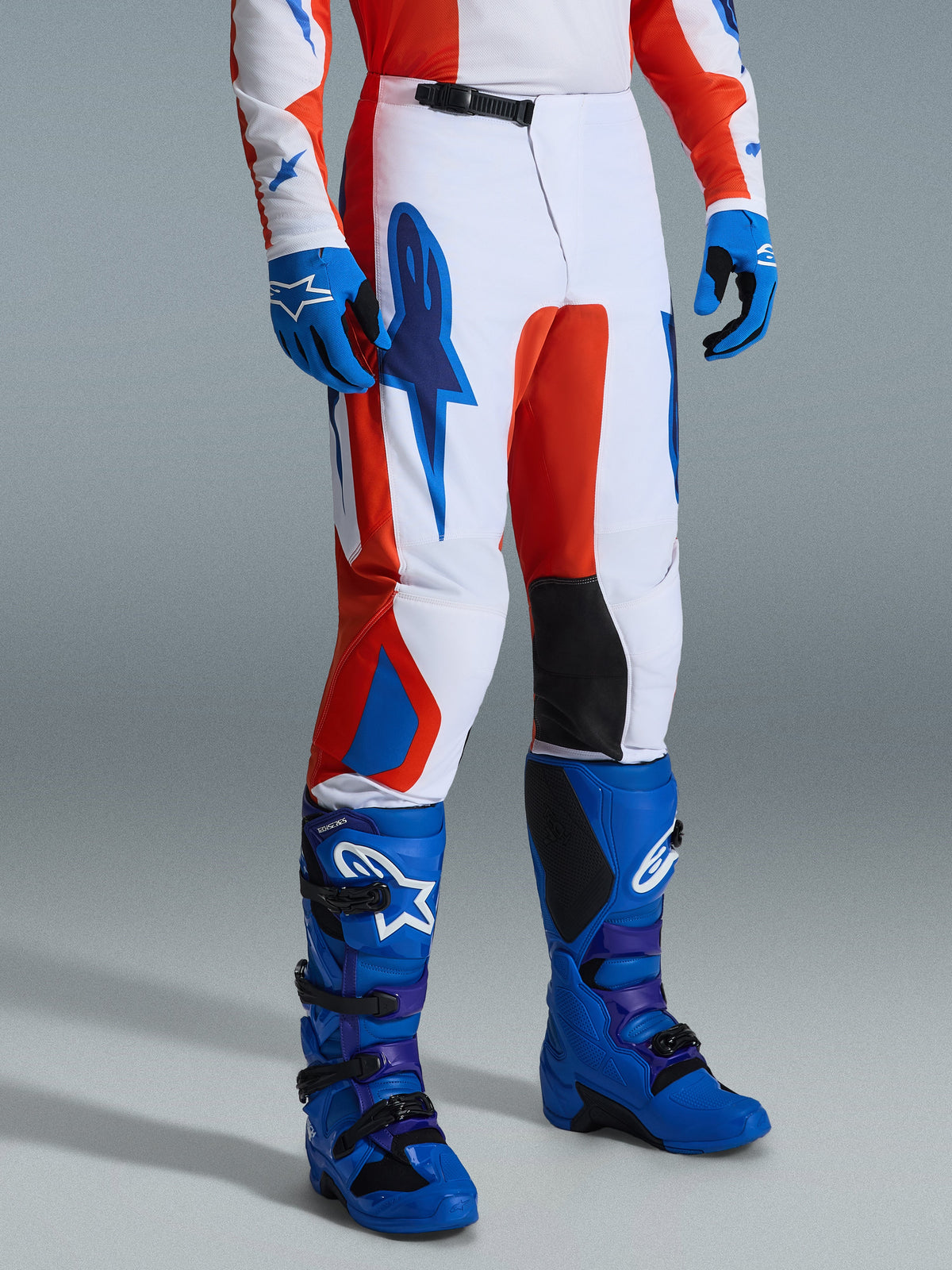 Alpinestars 2026 Fluid Grid byxor, motocrossbyxor, orange och UCLA-blå, bärs med matchande blå handskar och stövlar, vit bas med kraftfull grafisk design, svarta innerpaneler vid knäna, ergonomisk design för terrängkörning