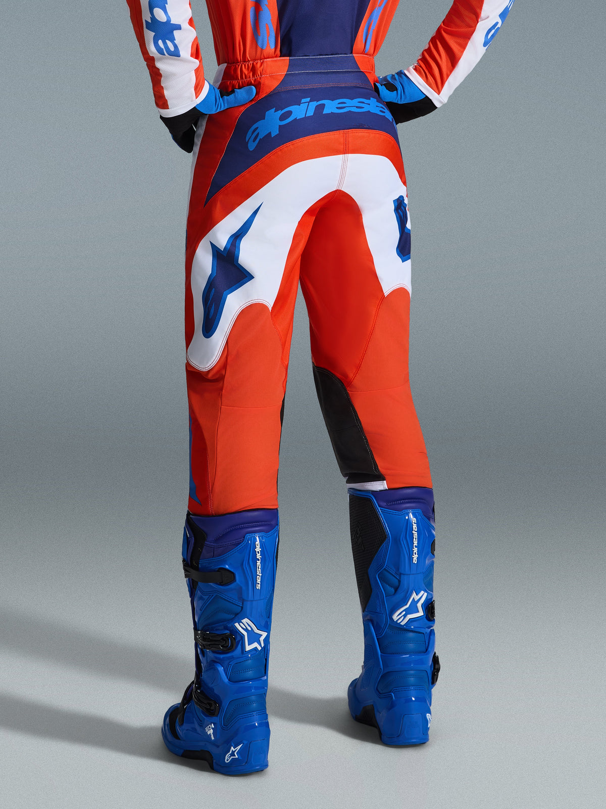 Alpinestars 2026 Fluid Grid byxor, motocrossbyxor, orange och UCLA-blå med vita paneler, vy bakifrån som visar framträdande blå logotyp på sätet och stjärnlogotyp, matchade med blå motocross-stövlar, professionell utrustning för terrängkörning