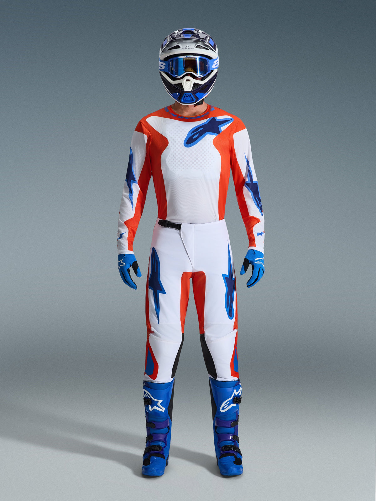 Motocrossförare i komplett racingutrustning, Alpinestars 2026 Fluid Grid-byxor i Orange och UCLA Blue, vit tröja med orangea ärmar och blå logotyper, matchande blå hjälm och goggles, blå stövlar och blå handskar, koordinerad outfit för terrängkörning