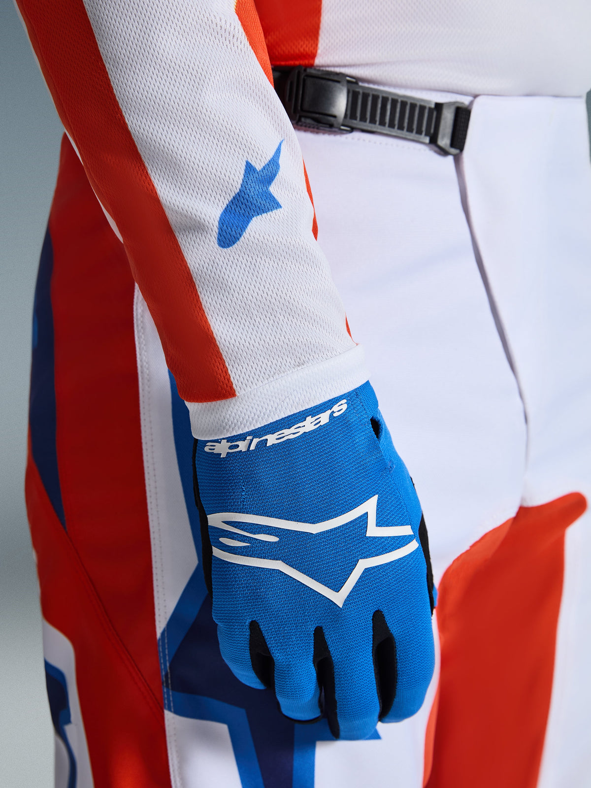 Alpinestars 2026 Fluid Grid byxor, motocrossbyxor, orange och UCLA-blå, närbild med matchande blå handske och vit tröjärm i mesh med blå logotyper, visar spärrspännet i midjan och atletisk passform