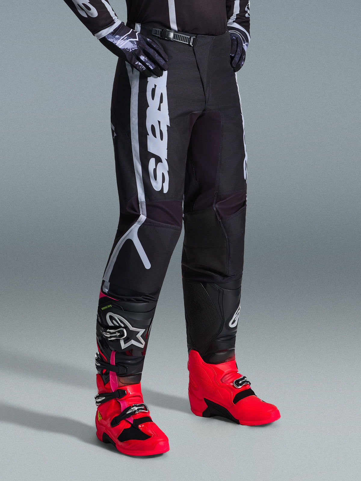 Alpinestars 2026 Fluid Apex byxor, motocrossbyxor, svarta och grå, bärs med färgstarka rosa och röda stövlar och matchande svartvit tröja, med framträdande vertikal Alpinestars-logotyp på låret, förstärkta paneler och spärrspänne i midjan för terrängprestanda.