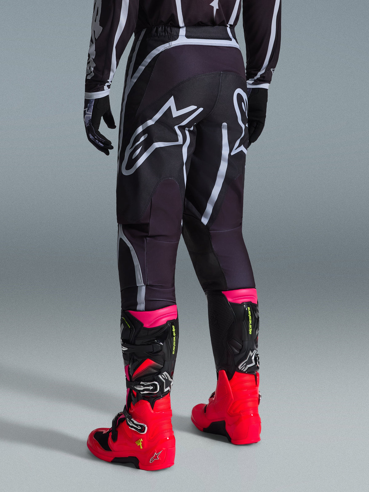 Alpinestars 2026 Fluid Apex byxor, motocrossbyxor, svarta och grå, vy bakifrån med stora vita Alpinestars-logotyper på sätet, bärs med starkt röda och rosa stövlar, hållbar 600D-polytygskonstruktion för terrängprestanda