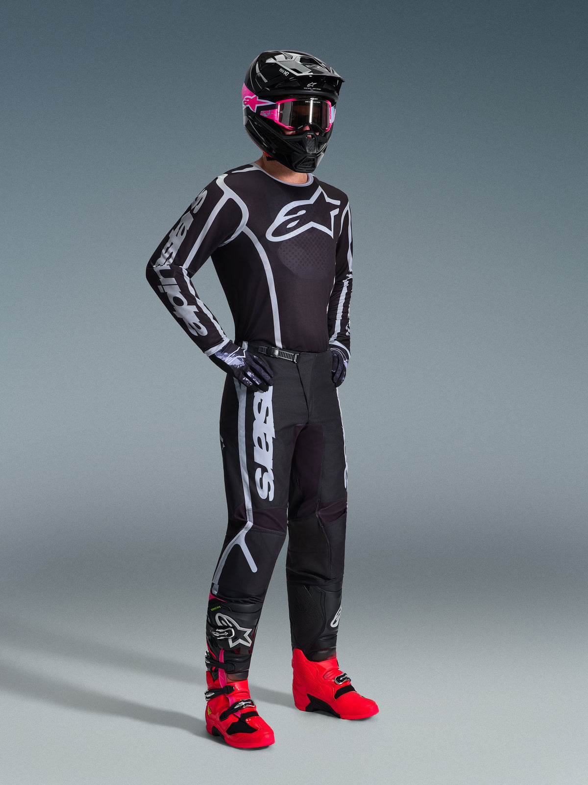 Alpinestars 2026 Fluid Apex byxor, motocrossbyxor, svarta och grå, bärs med matchande svart och vit tröja, svart hjälm med rosa goggles och klarröda stövlar, med tydlig vit logotyp på benen och sportig passform för terrängkörning.