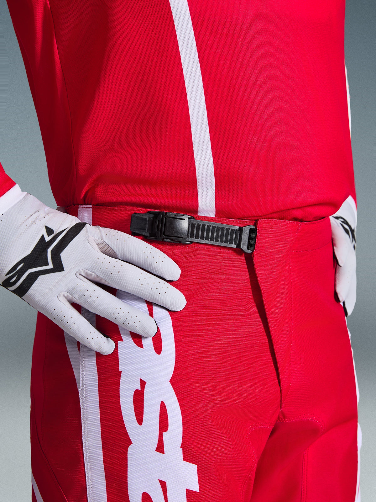 Alpinestars 2026 Fluid Apex byxor, motocrossbyxor, röda och vita, närbild på det svarta spännet med spärr i midjan, bärs med vita Alpinestars-handskar och matchande röd tröja, slitstark prestandautrustning för terrängkörning