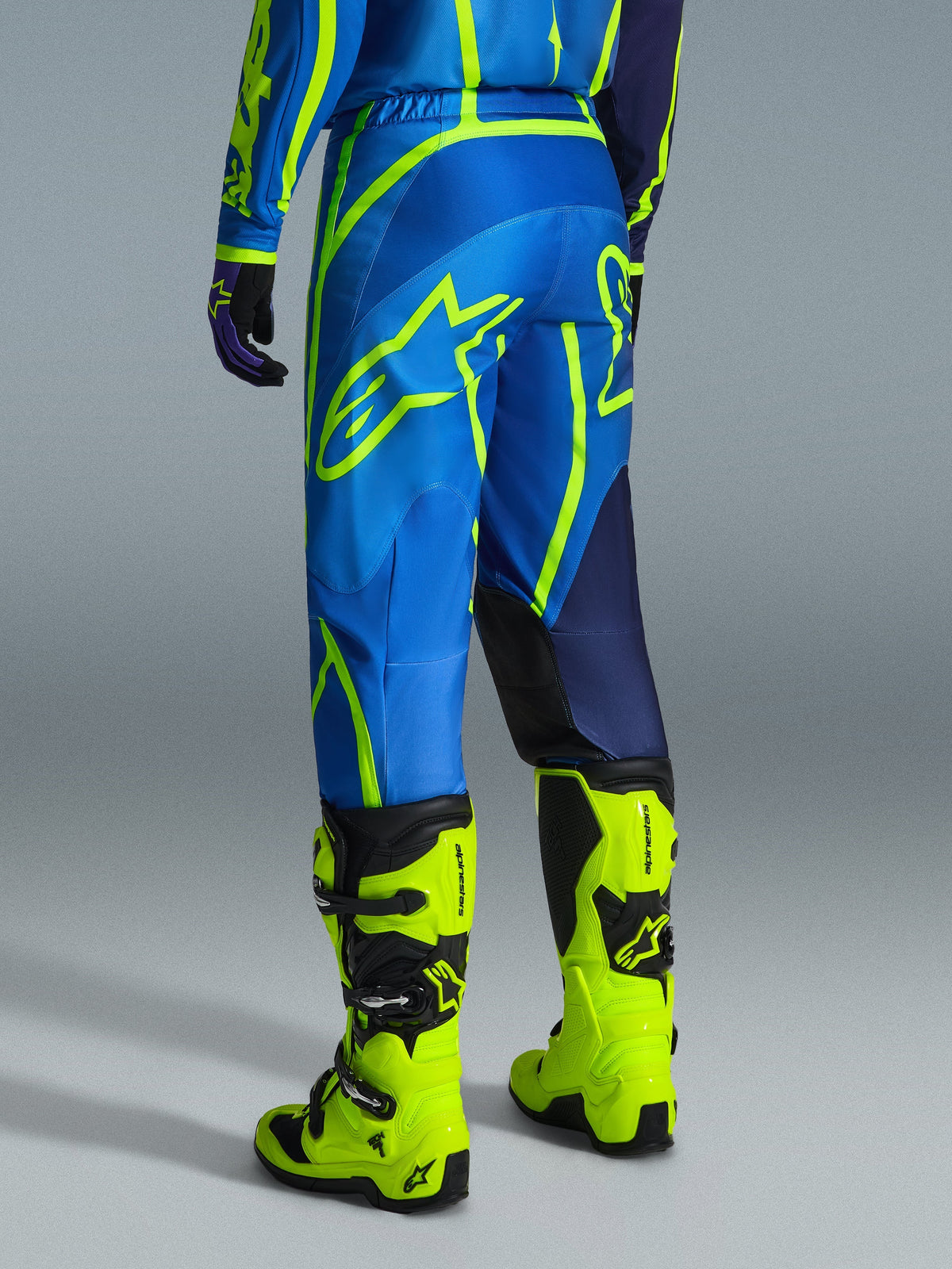 Alpinestars 2026 Fluid Apex byxor, motocrossbyxor, blå och neongula, vy bakifrån av förare i matchande utrustning och neongula stövlar, med framträdande Alpinestars-logotyper och ergonomisk design