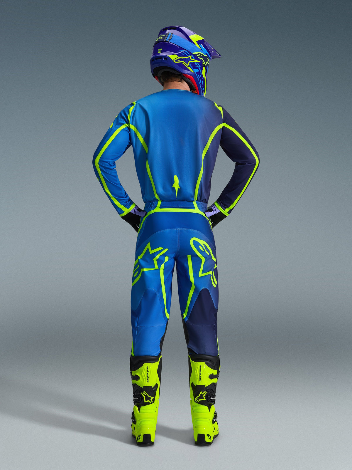 Alpinestars 2026 Fluid Apex-byxor, motocrossbyxor, blå och gul fluo, vy bakifrån av förare i full tävlingsutrustning med matchande tröja och hjälm, klargula stövlar, atletisk passform för motocross.