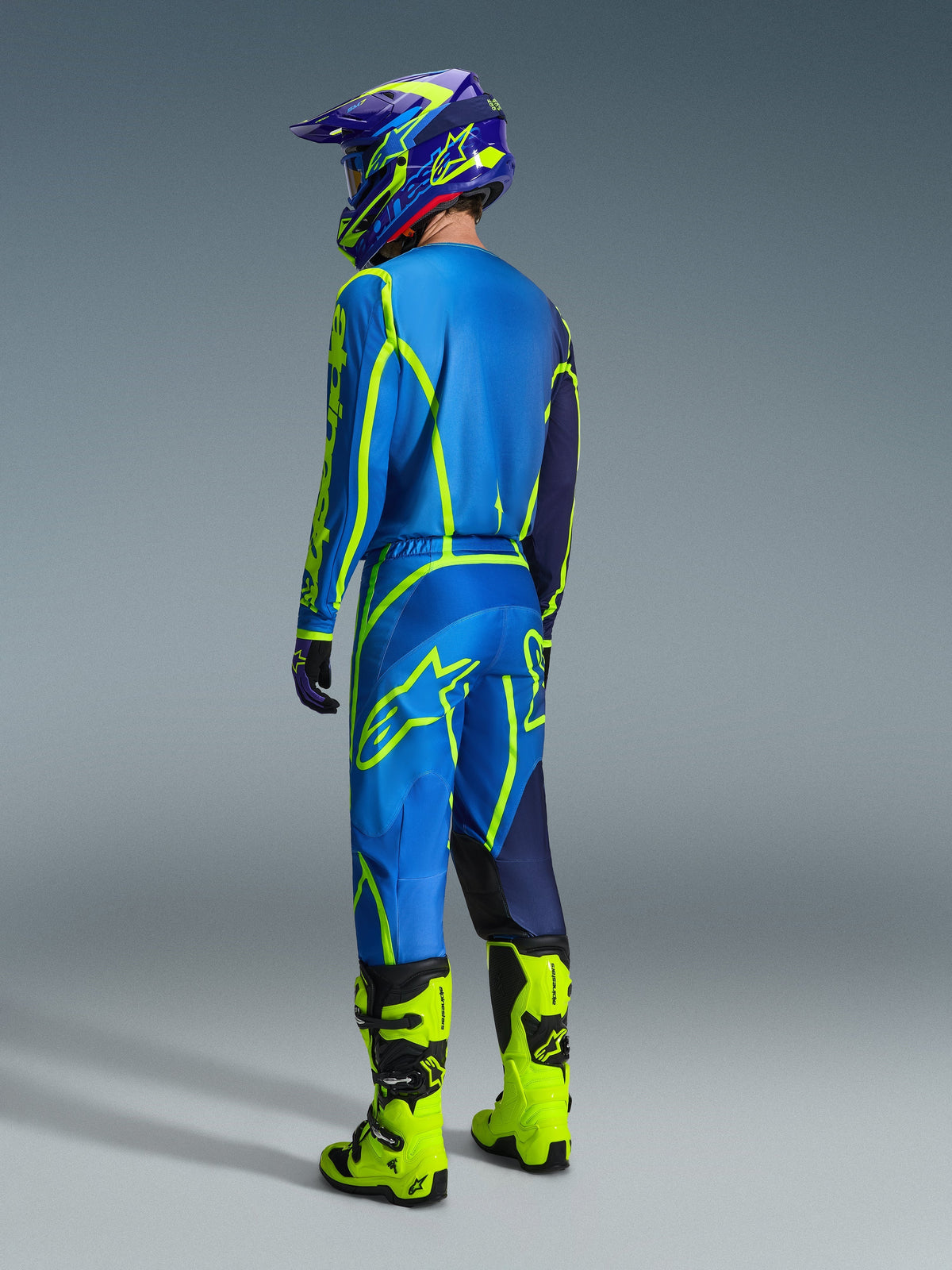 Motocrossförare sedd bakifrån bärande 2026 Alpinestars Fluid Apex-byxor i blått och gult fluo, matchande blå och gul tröja och hjälm, samt klargula neonstövlar, koordinerad racingutrustning för offroad-prestanda.