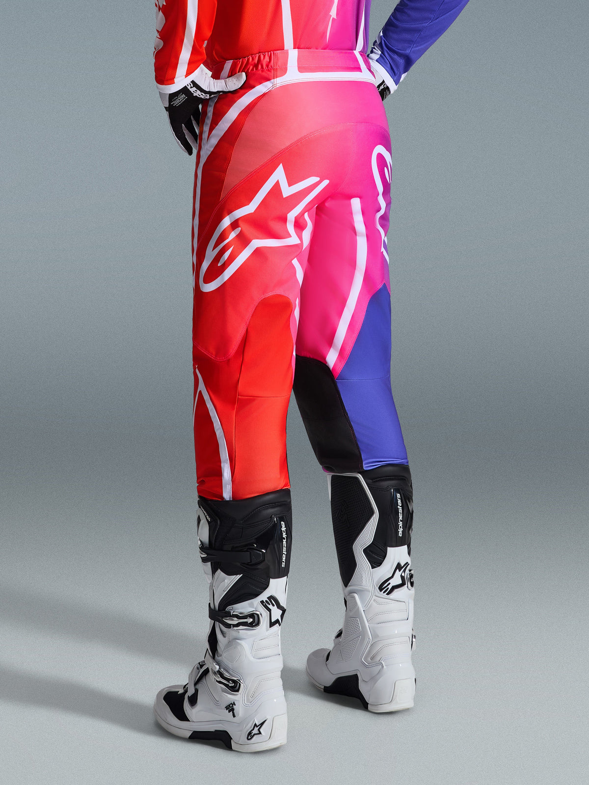 Alpinestars 2026 Fluid Apex-byxor, motocrossbyxor, flerfärgade i rött, rosa och lila, vy bakifrån av förare som bär byxor med vita och svarta motocross-stövlar, stor vit Alpinestars-logotyp, livfull tonad design