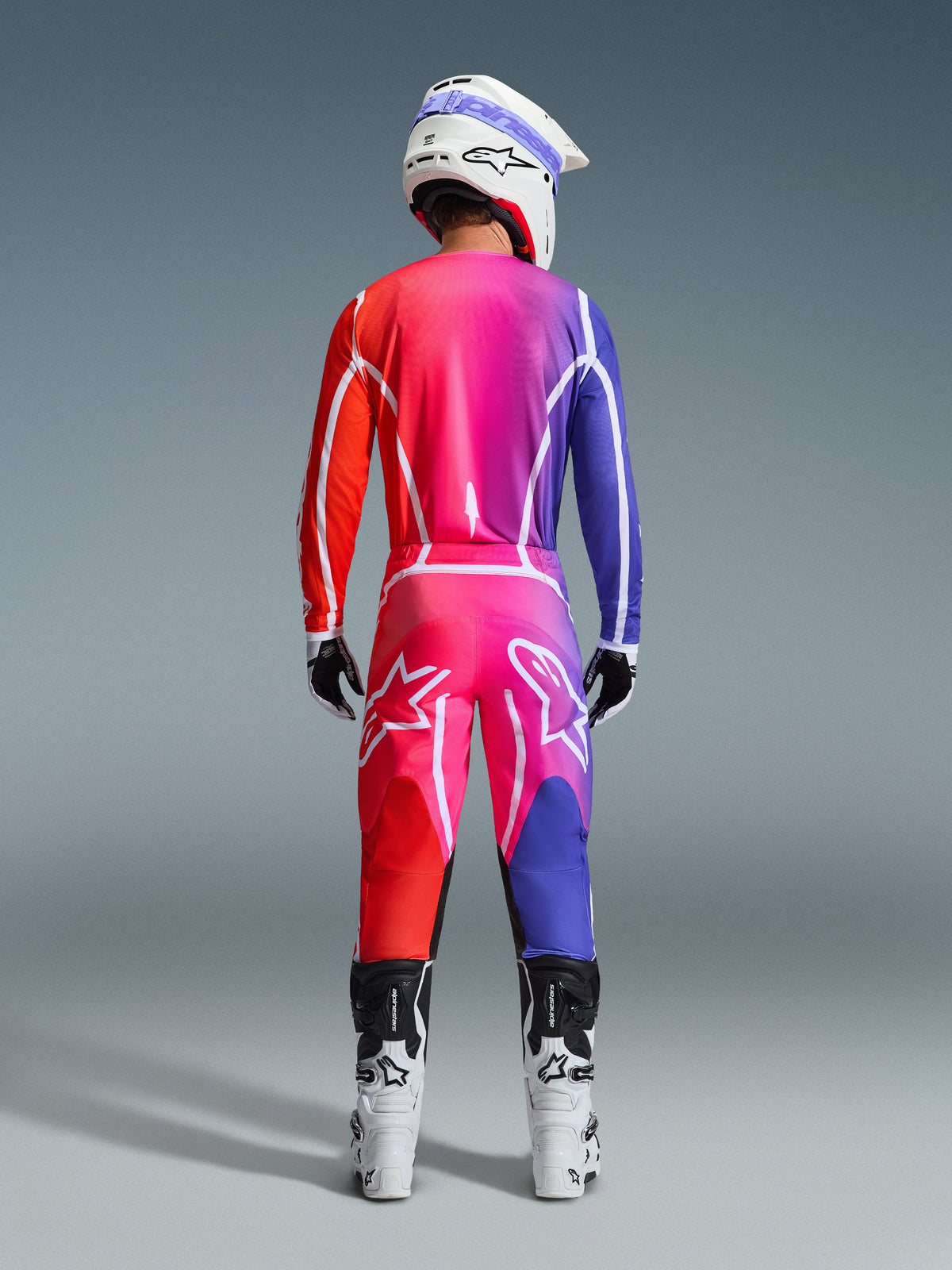 Motocrossförare sedd bakifrån i Alpinestars 2026 Fluid Apex-byxor och matchande tröja med en livfull gradient i rött, rosa och lila, kombinerat med vit hjälm och vita stövlar, komplett koordinerad utrustning för terrängkörning