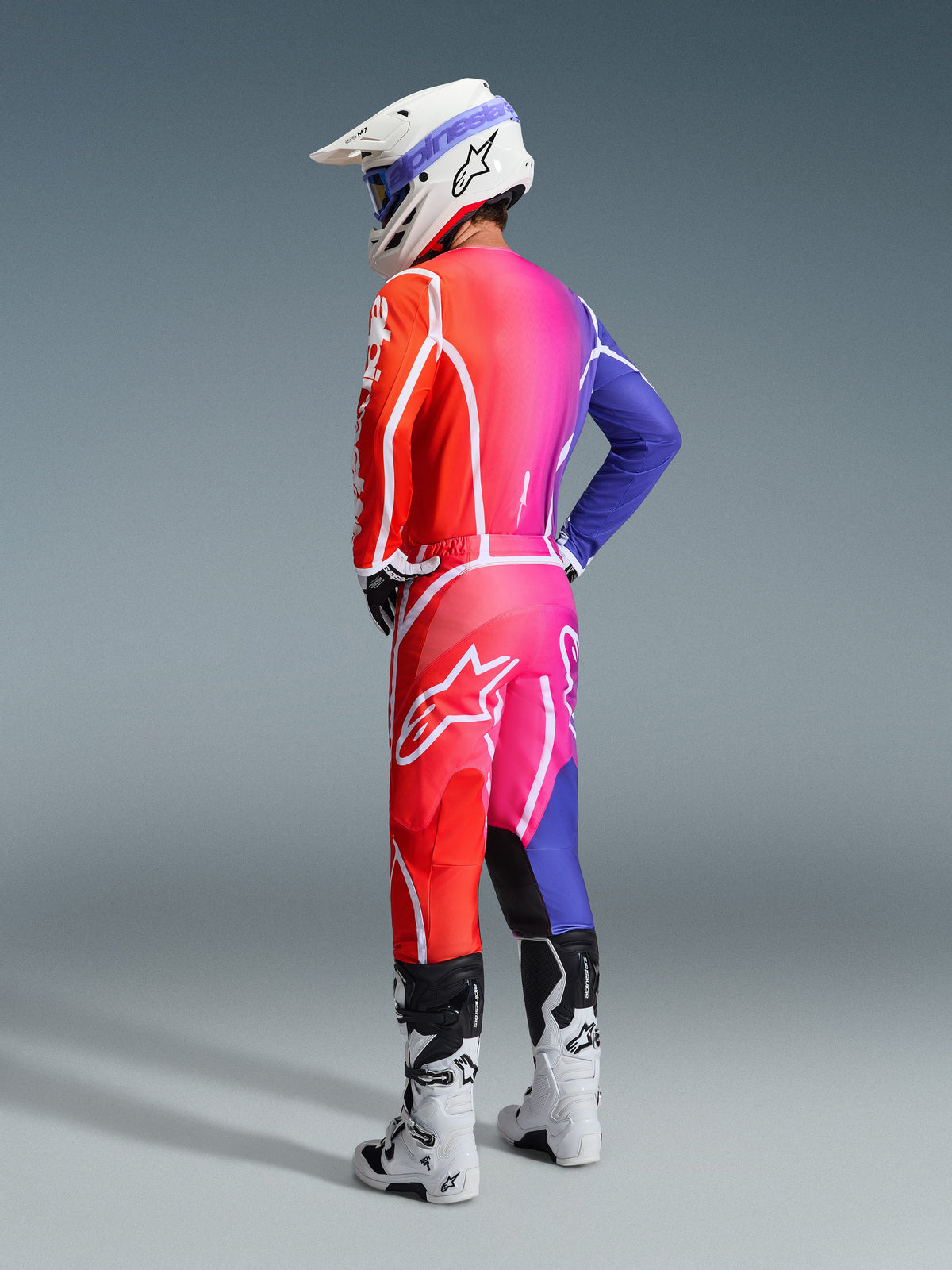 Motocrossförare i Alpinestars 2026 Fluid Apex-utrustning, flerfärgad tröja och byxor med röd, rosa och lila färgtoning och vita logotyper, vit hjälm och vita stövlar, koordinerad outfit för terrängkörning