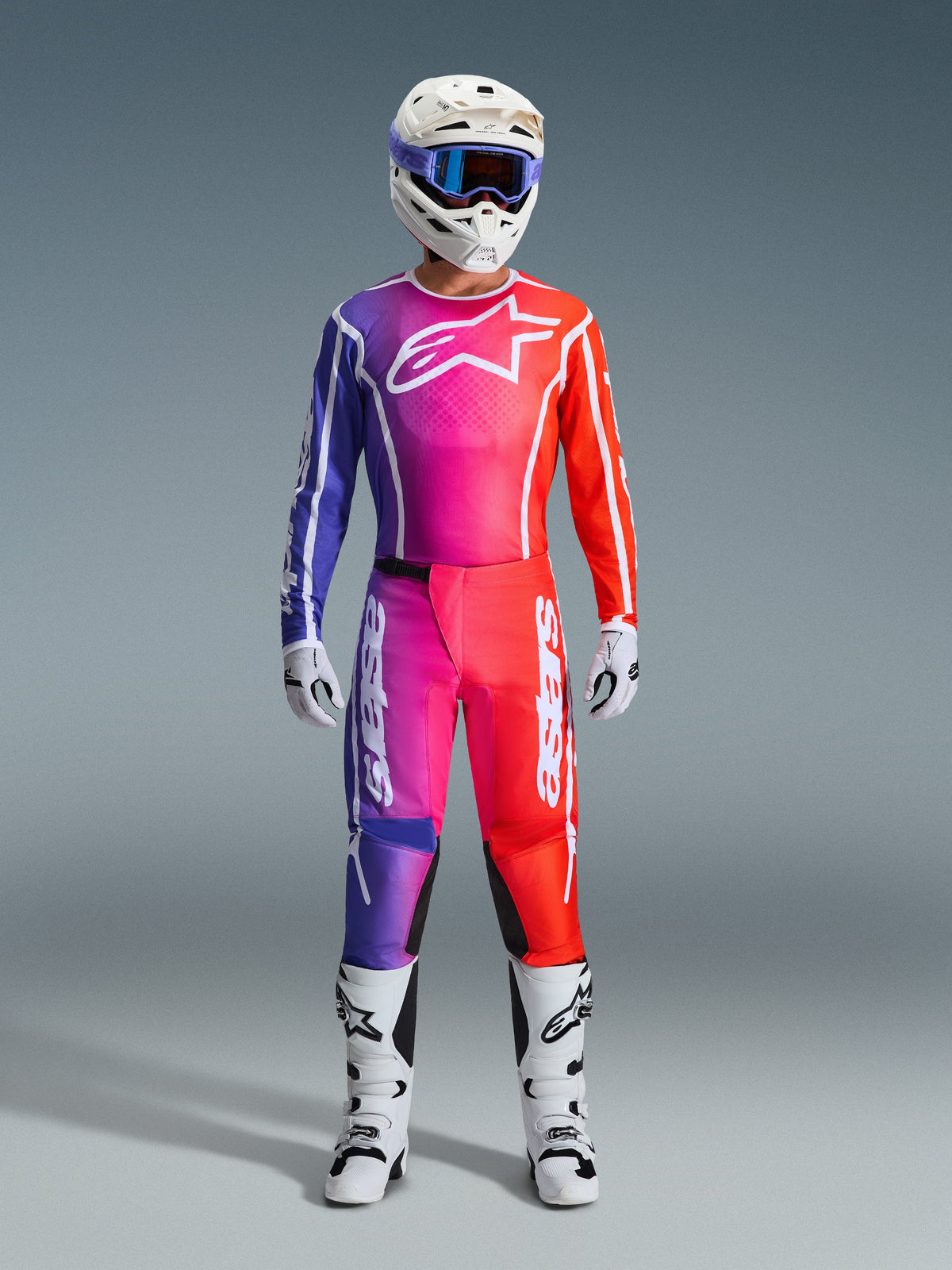 Motocrossförare iklädd 2026 Fluid Apex-byxor och matchande tröja, lila och orange färgtoning, vit hjälm med goggles, vita stövlar och handskar, koordinerad Alpinestars-racingutrustning för offroad-prestanda.