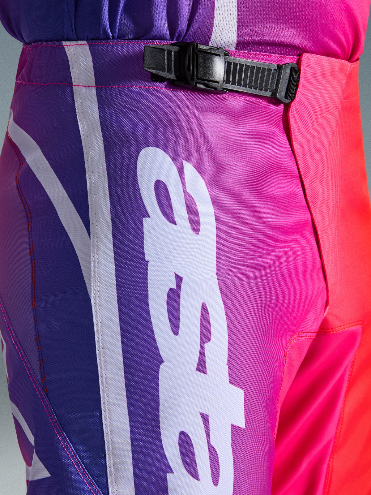 Alpinestars 2026 Fluid Apex byxor, motocrossbyxor, flerfärgade i lila, rosa och rött, närbild på midje- och höftpartiet med ett svart spänne med spärr och en stor vit vertikal Alpinestars-logotyp, livfull gradientdesign med slitstark textur i polytyg