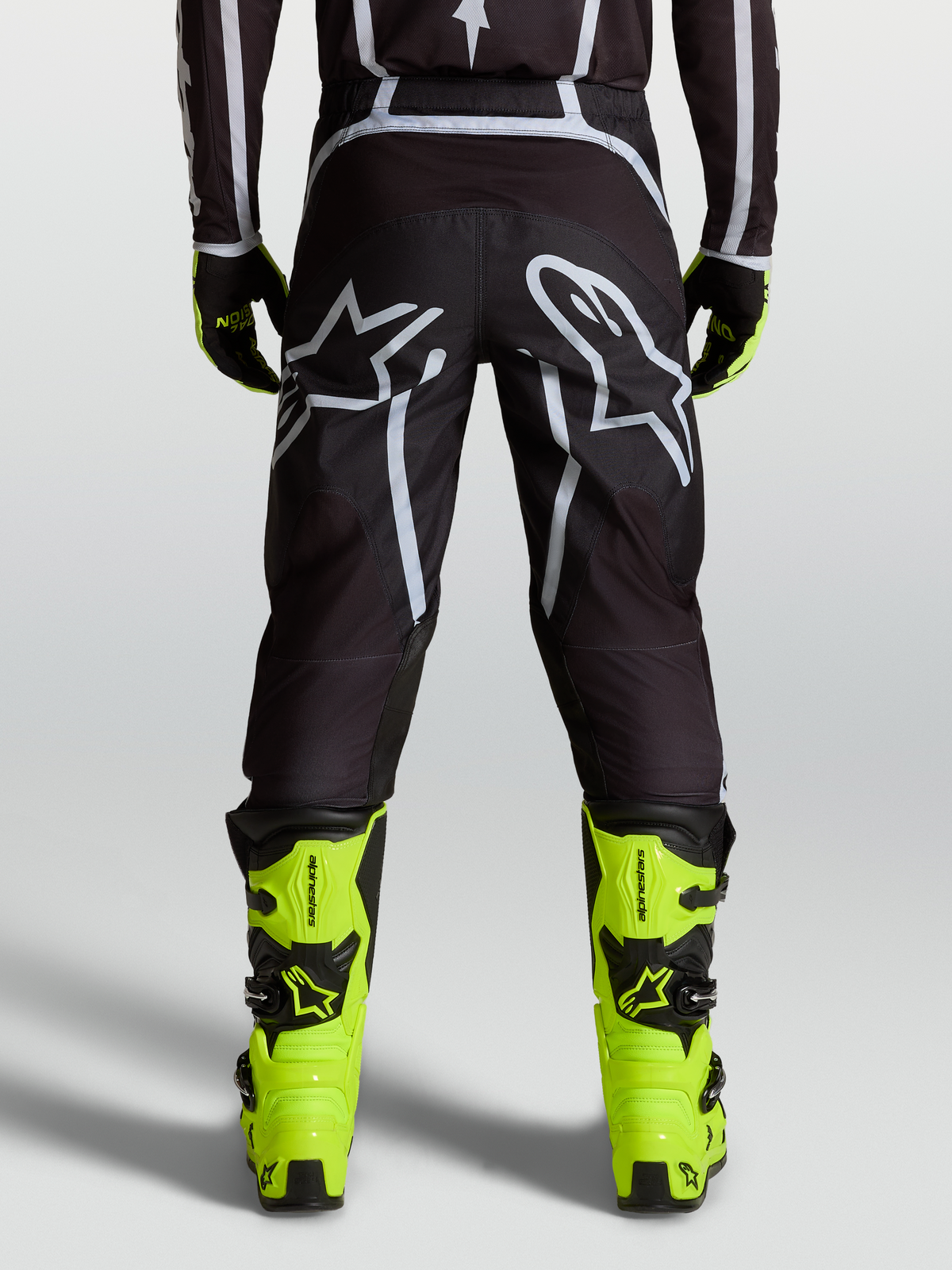 Alpinestars Fluid Apex V2 byxor, svarta och silverfärgade motocrossbyxor för herrar, sedda bakifrån på modell med neongula stövlar, ergonomisk passform och stor Astars-logotyp.