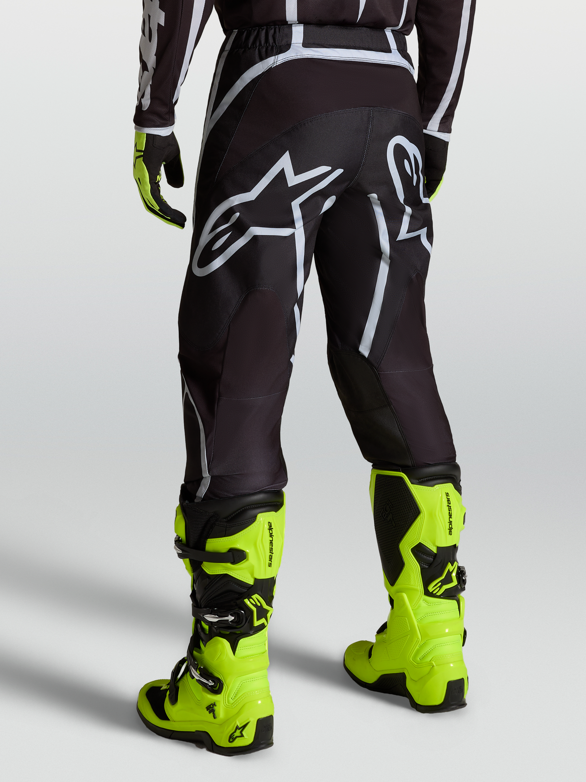 Alpinestars Fluid Apex V2 byxor, motocrossbyxor för herr i svart och silver, sedda bakifrån med neongula stövlar.