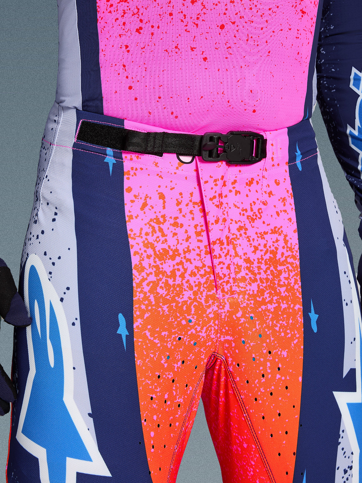 Alpinestars Limited Edition Supertech Records byxor, motocrossbyxor, flerfärgade i rosa och orange med mörkblå sidopaneler, närbild av midjan med ett svart SX1 självlåsande magnetspänne, rip-stop-tyg med stänkgrafik och laserskurna perforeringar.
