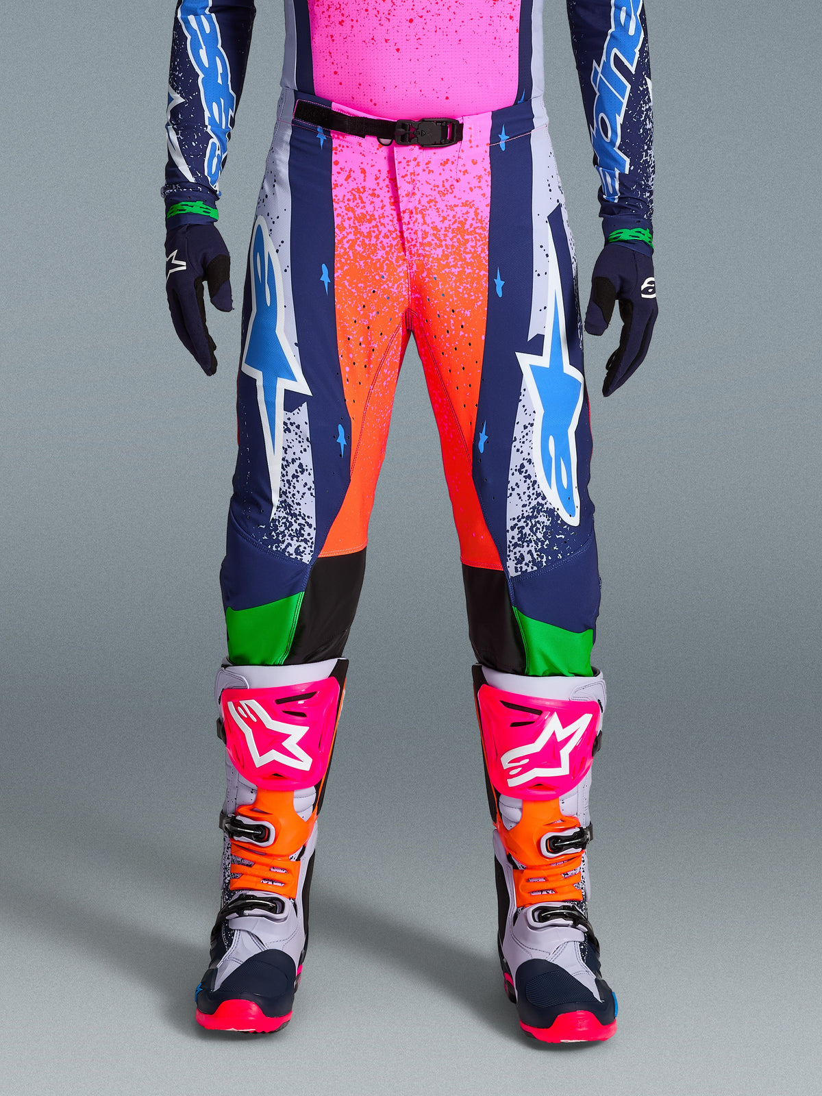 Alpinestars Limited Edition Supertech Records byxor, motocrossbyxor, flerfärgade med livfull rosa och orange splash-grafik, marinblå paneler med blå stjärnlogotyper och gröna detaljer. Bärs med matchande tröja och grå, rosa och orangea motocross-stövlar.