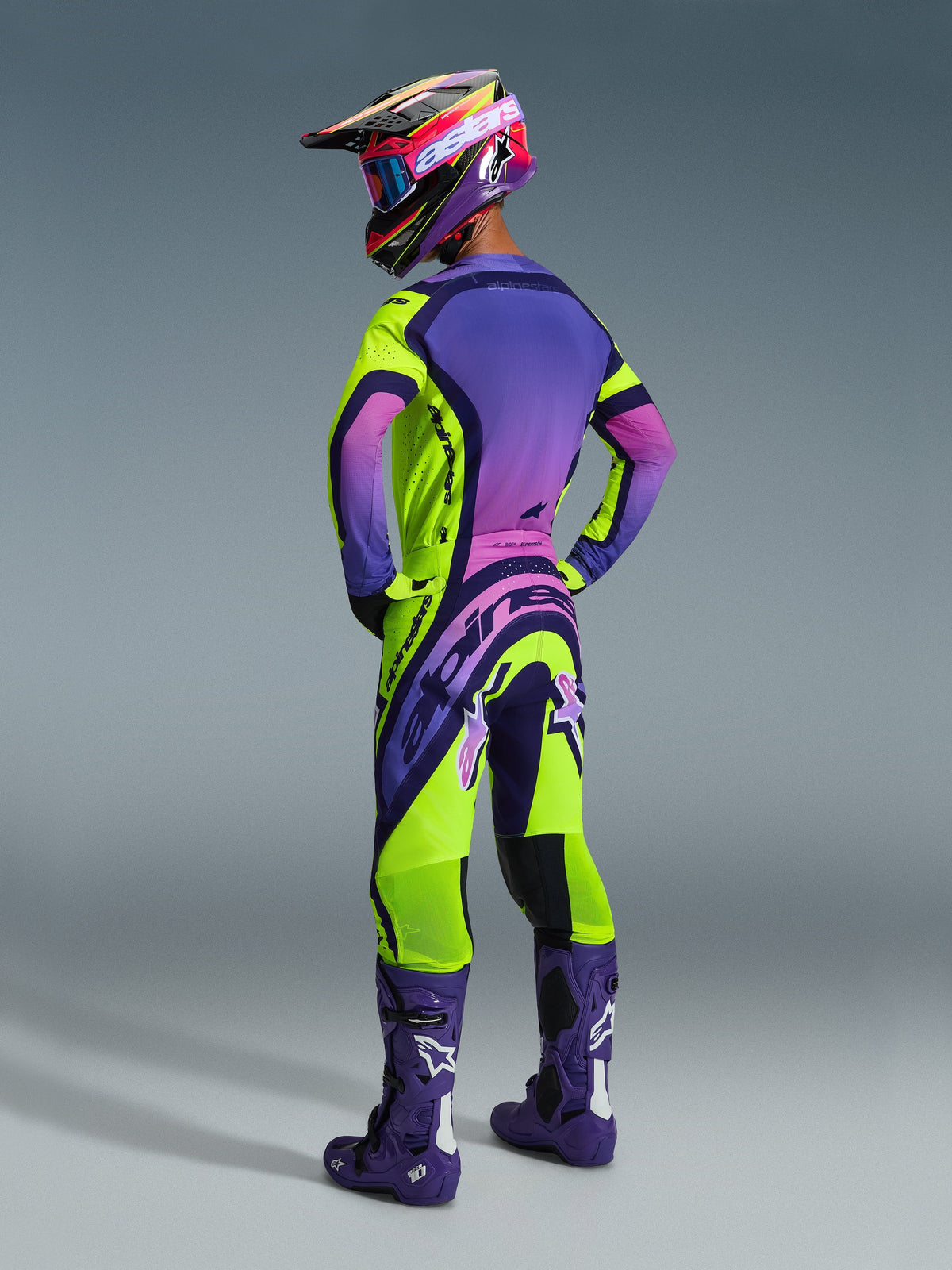 Motocross rider wearing complete racing gear from the back, 2026 Supertech Vista Jersey in Yellow Purple and Black, matching pants with bold branding, purple helmet with pink goggles, and purple boots with white logos, coordinated professional motocross outfit.