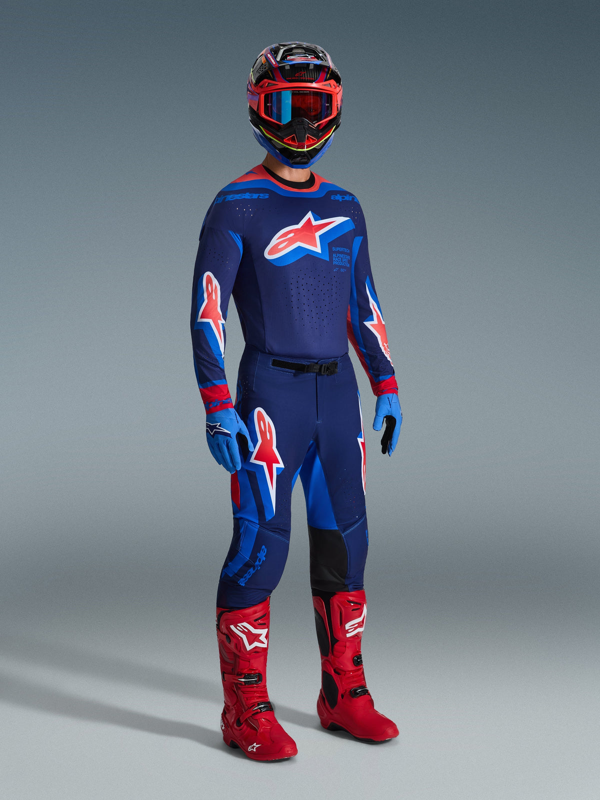 Motocross rider wearing 2026 Supertech Vista Pants in Dark Navy Hot Coral Cobalt, matching long-sleeve racing jersey, red motocross boots, and coordinated helmet with red goggles, full-body studio shot of high-performance off-road gear