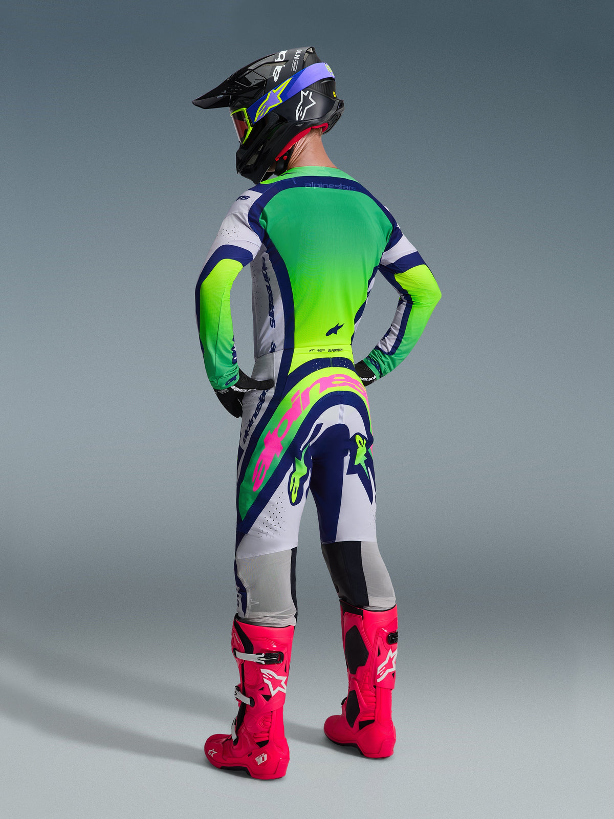 Rear view of a motocross rider wearing the Alpinestars 2026 Supertech Vista Gear Set, white and lime green jersey with blue accents, matching pants with a bold pink logo across the back, bright pink racing boots, and a black helmet with lime green goggles, professional off-road racing apparel