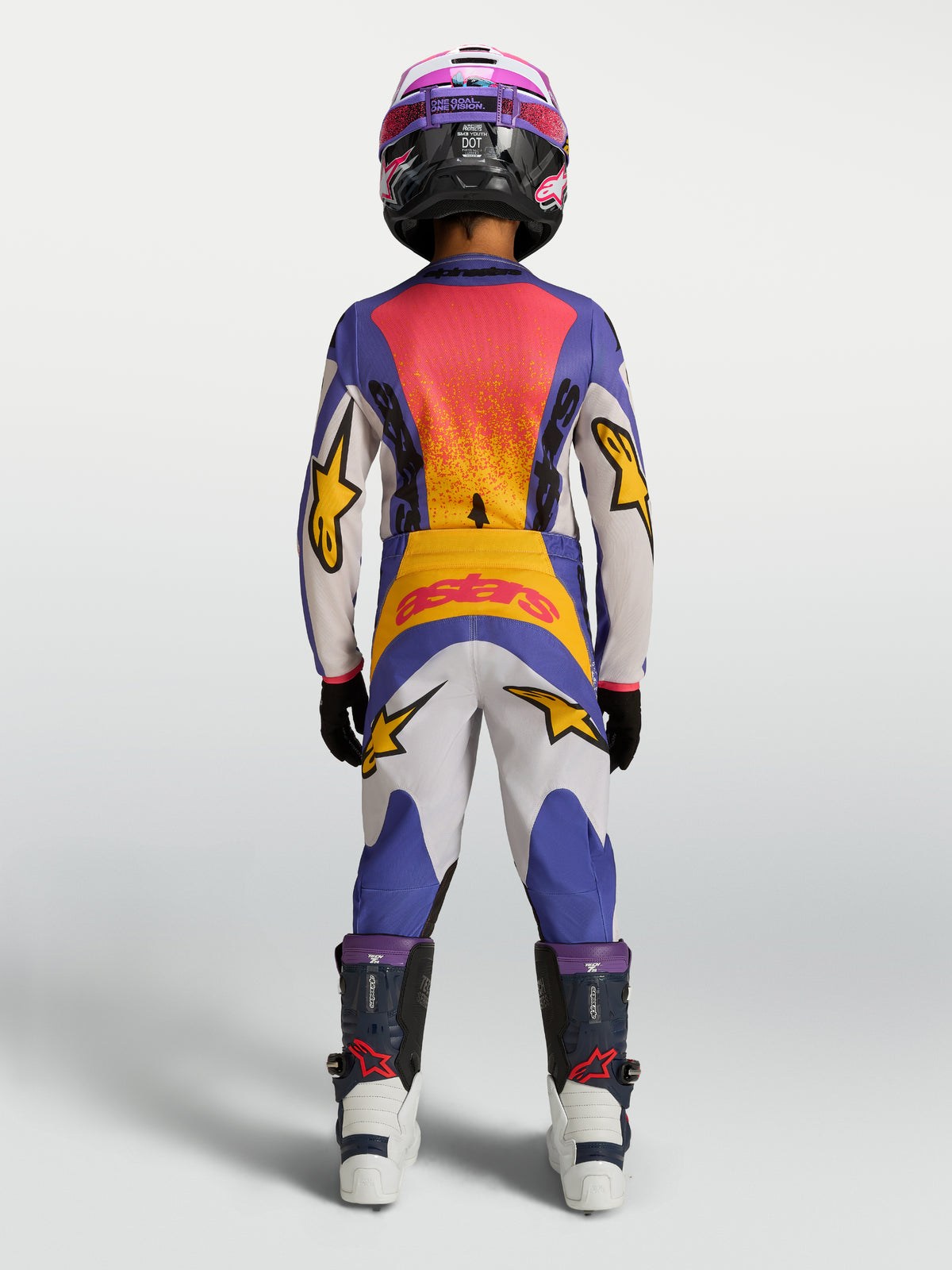 Alpinestars Ungdom Fluid Drag byxor, motocrossbyxor för terrängkörning, blåa och vita med gula accenter, sedda bakifrån på en ung förare med matchande tröja och hjälm.
