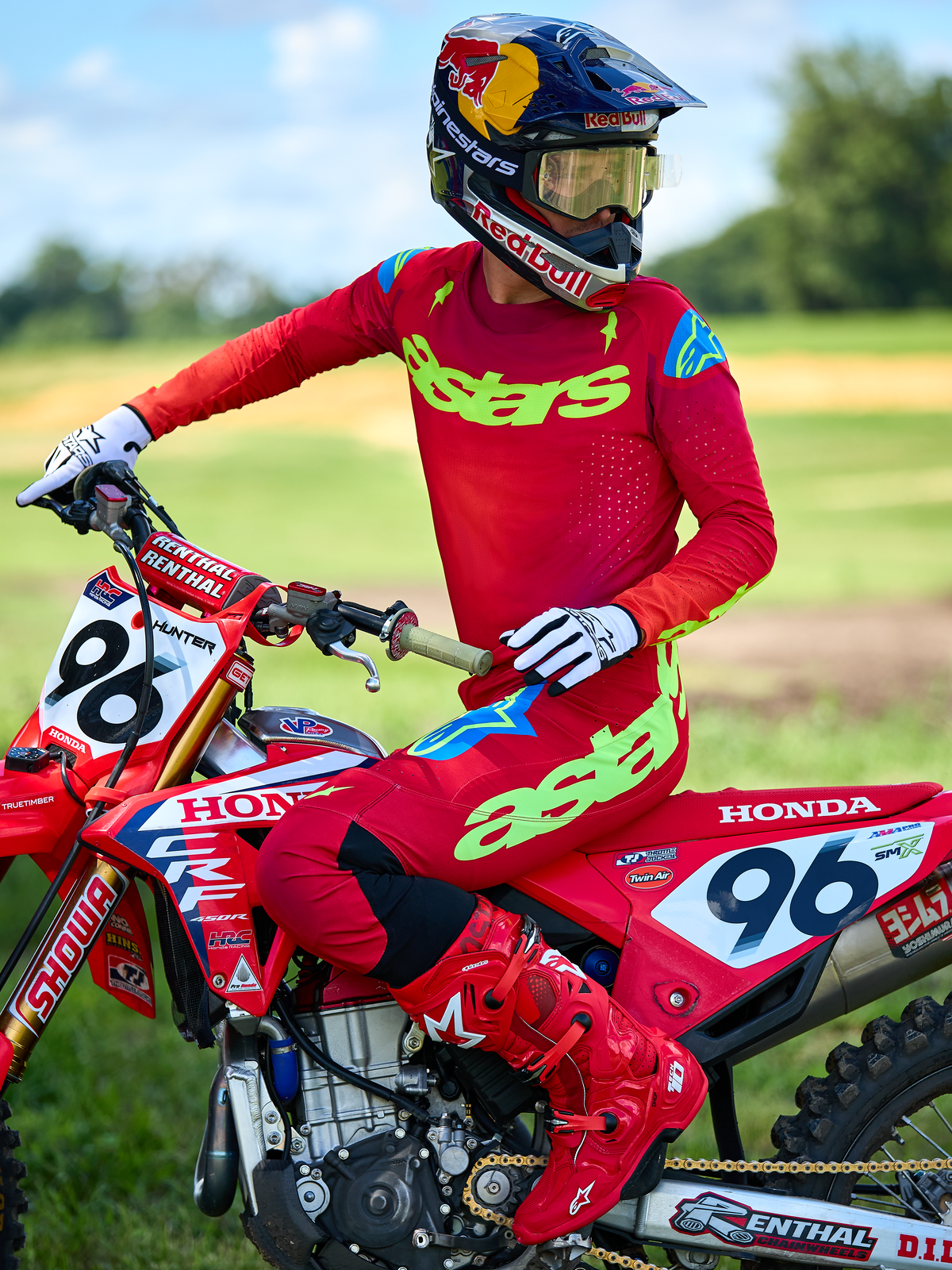 Motocrossförare på en röd Honda-motorcykel iklädd Alpinestars 2025 Supertech Maker-utrustningsset i rött, med en röd tröja med neongrön 'astars'-logotyp och blå detaljer på axlarna, matchande röda byxor, helröda stövlar, vita handskar och en Red Bull-hjälm, professionellt tävlingsset för terrängkörning