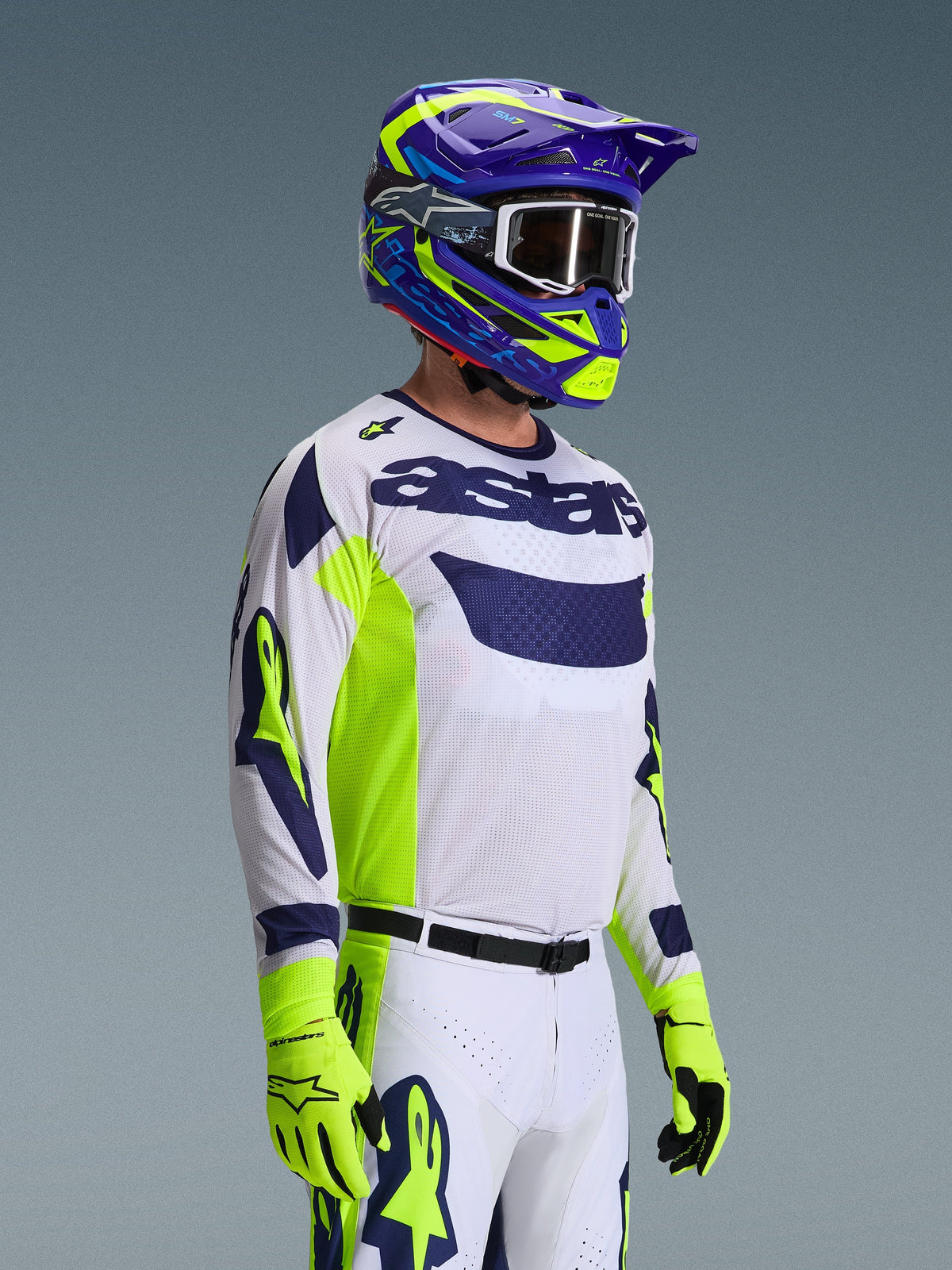 Motocrossförare i Alpinestars 2026 Racer Air Riway-utrustningsset, vit tröja med marinblå logotyp och neongula sidopaneler, vita byxor med matchande marinblå och gula detaljer, lila och neongul hjälm med vita goggles, neongula handskar, högpresterande terrängkörningsutrustning