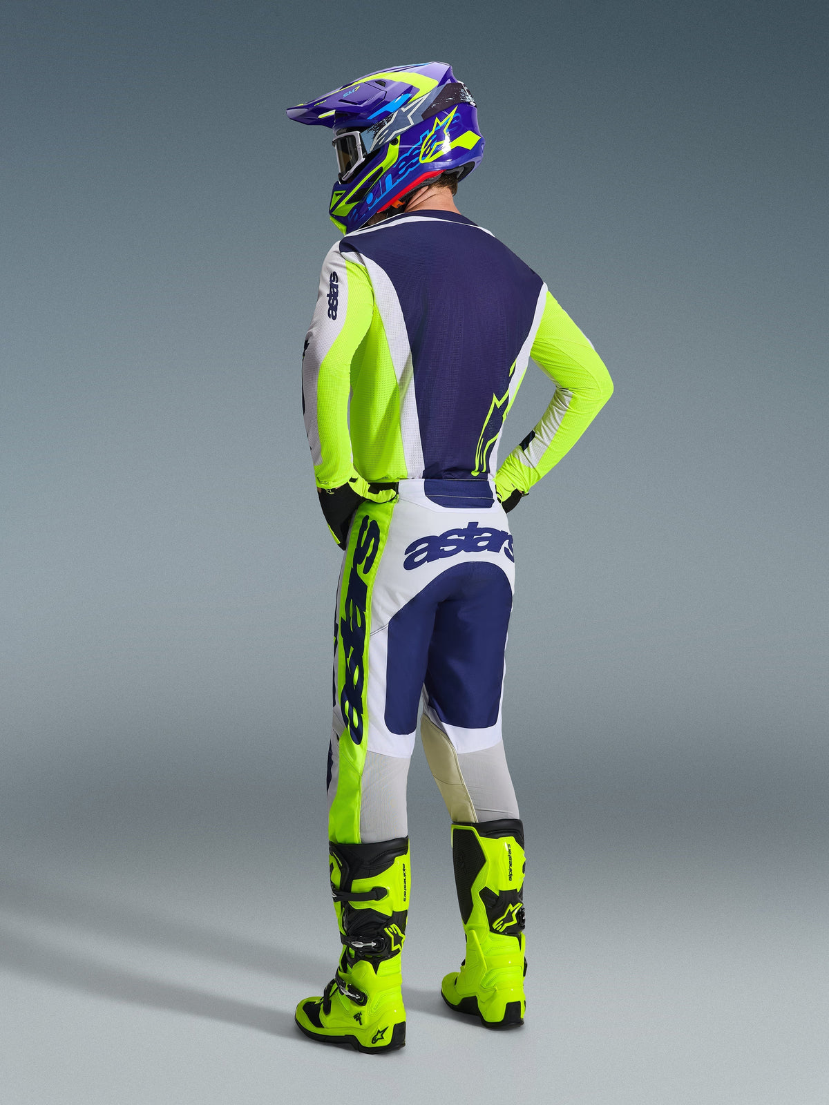 Motocrossförare sedd bakifrån iklädd Alpinestars 2026 Racer Air Riway utrustningsset, ljusgrå, gul fluo och blå tröja och byxor, med 'astars'-logotyper på ärmar och säte, neongula stövlar och en matchande färgstark hjälm.