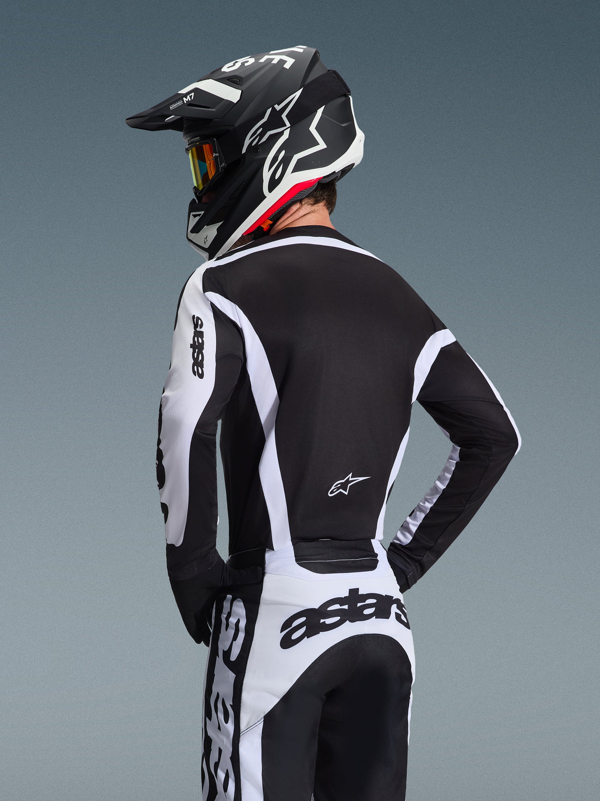 Motocrossförare sedd bakifrån i Alpinestars 2026 Racer Riway-tröja i vitt och svart, med 'astars'-logotyp på ärmarna och nedre delen av ryggen, matchande motocrossbyxor och en svart hjälm med goggles, samordnad utrustning för terrängkörning.